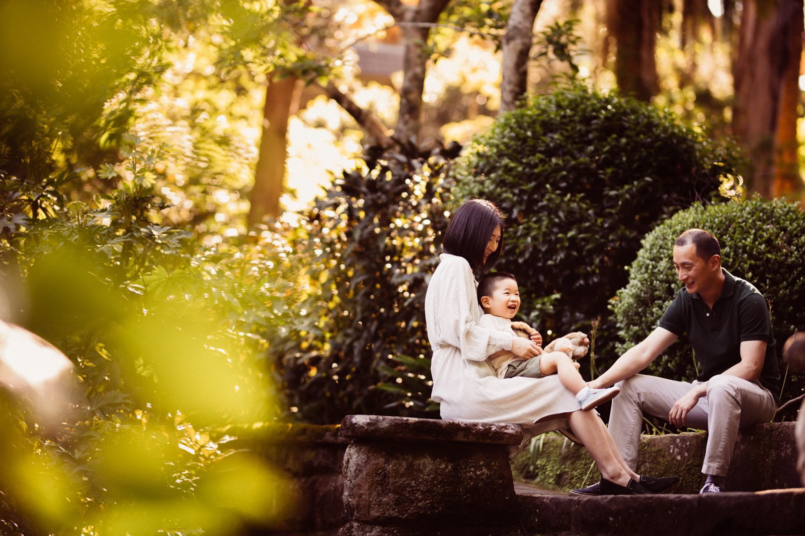 A family of three, including a woman, a man, and a young boy, sitting on a stone bench outdoors, surrounded by lush greenery, with sunlight filtering through trees. They are smiling and laughing, enjoying each other's company.