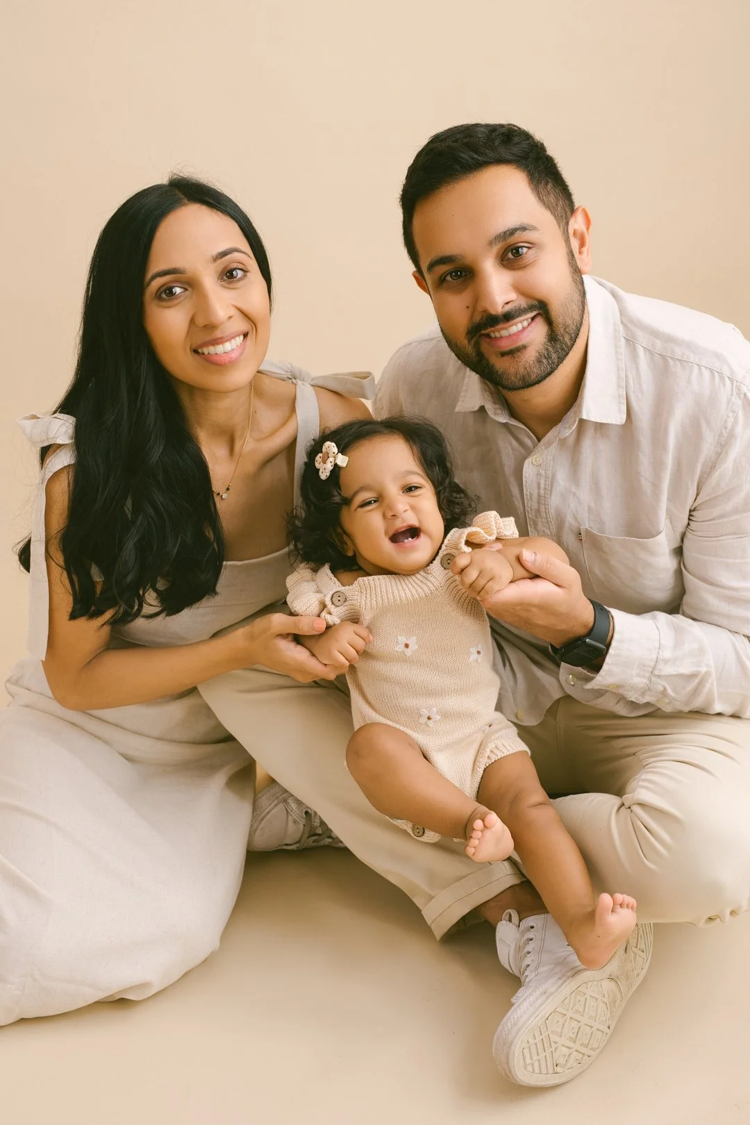 A happy family of three with a young girl, sitting on the floor against a beige background, smiling at the camera.