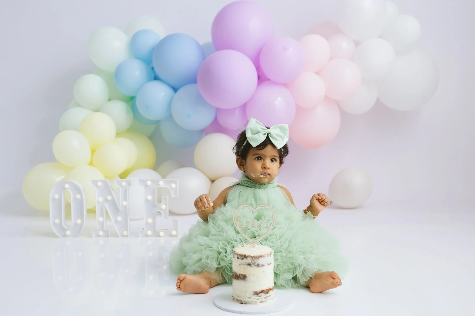 A A  one year old baby girl in a light green dress with a matching bow headband sits on the floor in front of a cake and pastel-colored balloons. The girl appears to be celebrating her first birthday, as indicated by the lit-up 