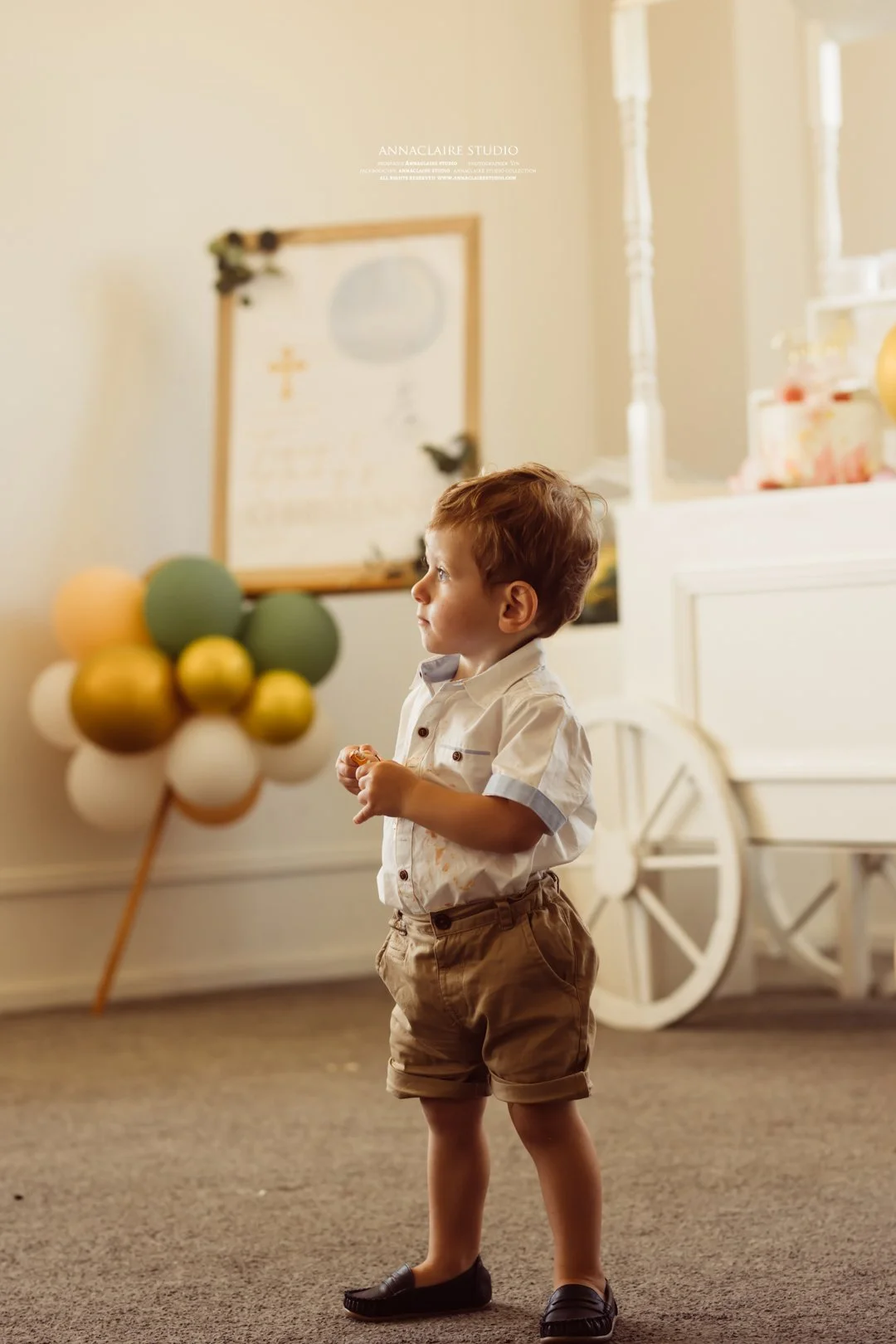 Young boy with curly hair wearing a white shirt and brown shorts standing indoors, holding a small object in his hand. In the background, there are balloons arranged in a cluster, a white cart with a large wheel, and a framed picture on the wall.