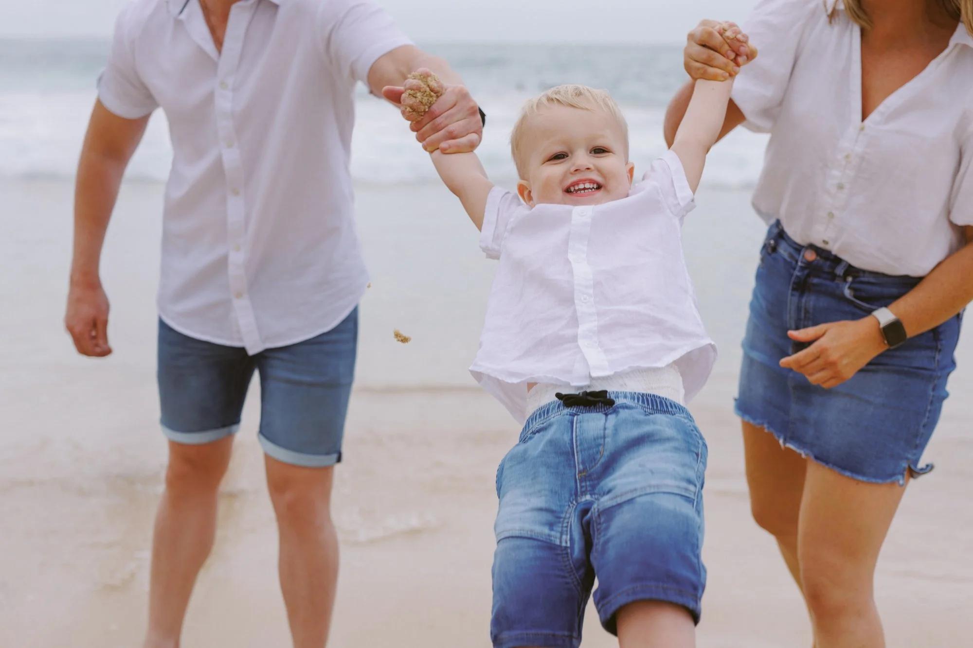 A young boy in a white shirt and blue shorts smiling while holding hands with two adults at the beach, with the ocean in the background.