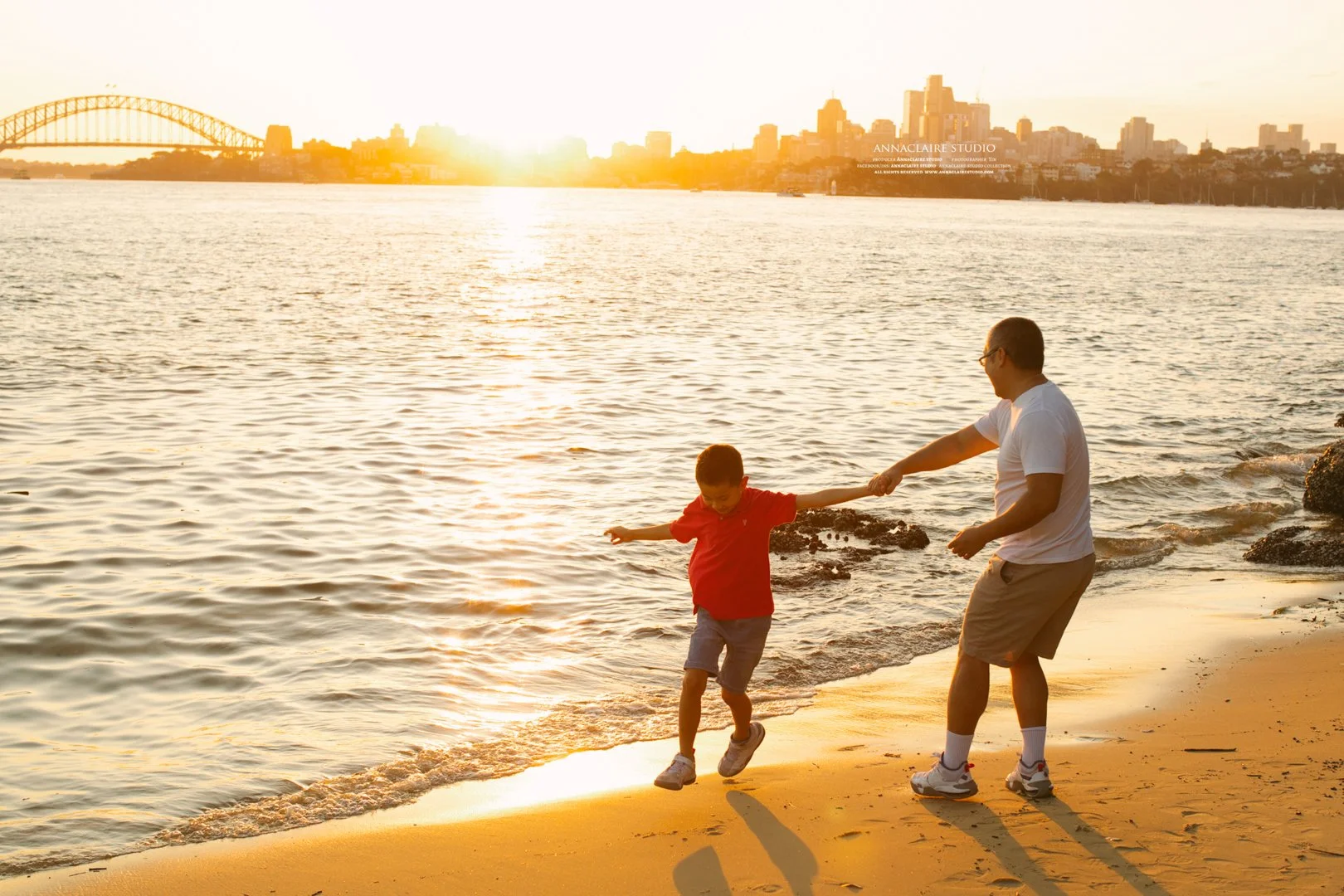 A father and son playing on the beach at sunset, with a Sydney city skyline and bridge in the background.