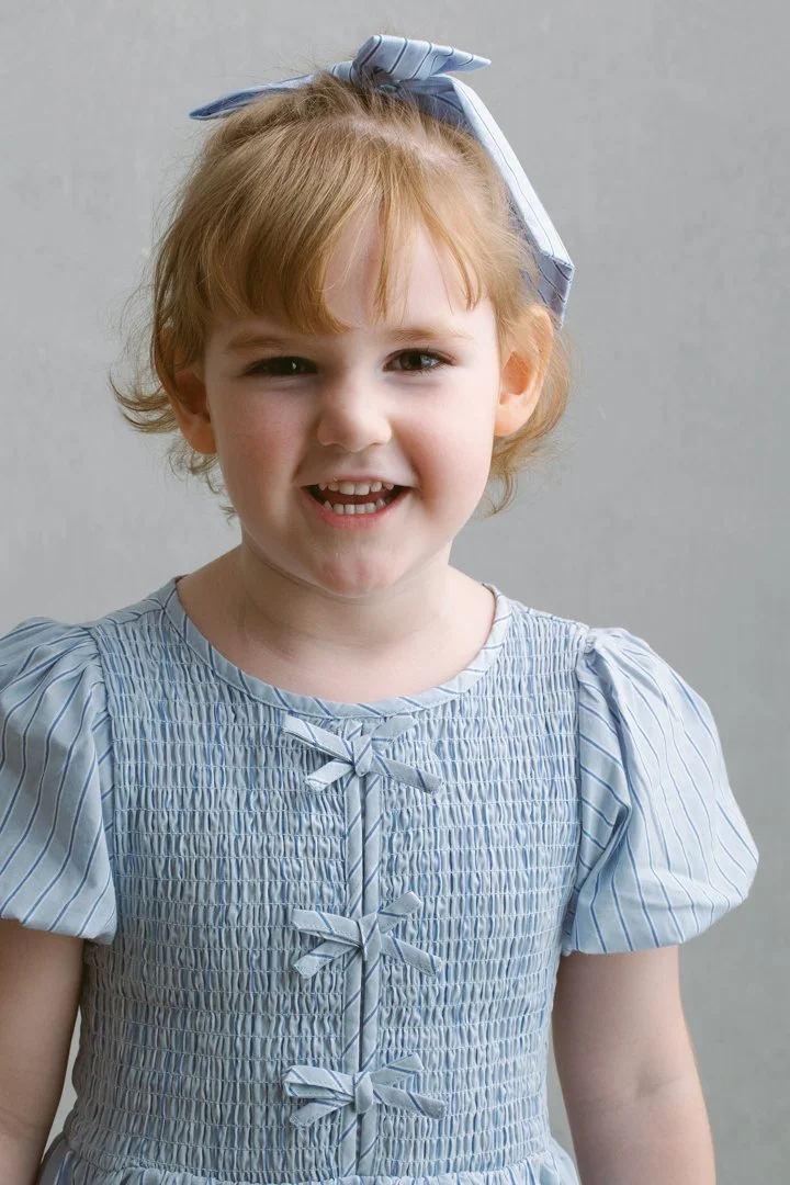 A young girl with red hair wearing a light blue dress with puffed sleeves and bow accents on the bodice, smiling at the camera, with a matching bow in her hair, against a plain gray background.