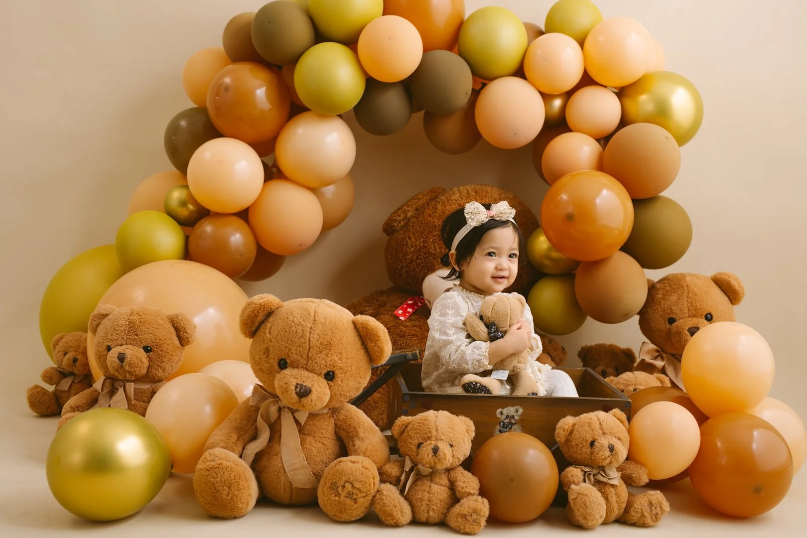 in this cake smash photo ,   1 year old baby  sitting in a wooden box surrounded by teddy bears with a balloon arch overhead. The bear arch is made of orange, green, beige, and gold balloons.