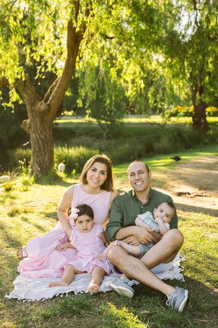 Family of four sitting on blanket in park, smiling at camera, surrounded by green trees and sunlight in centennial PARK .