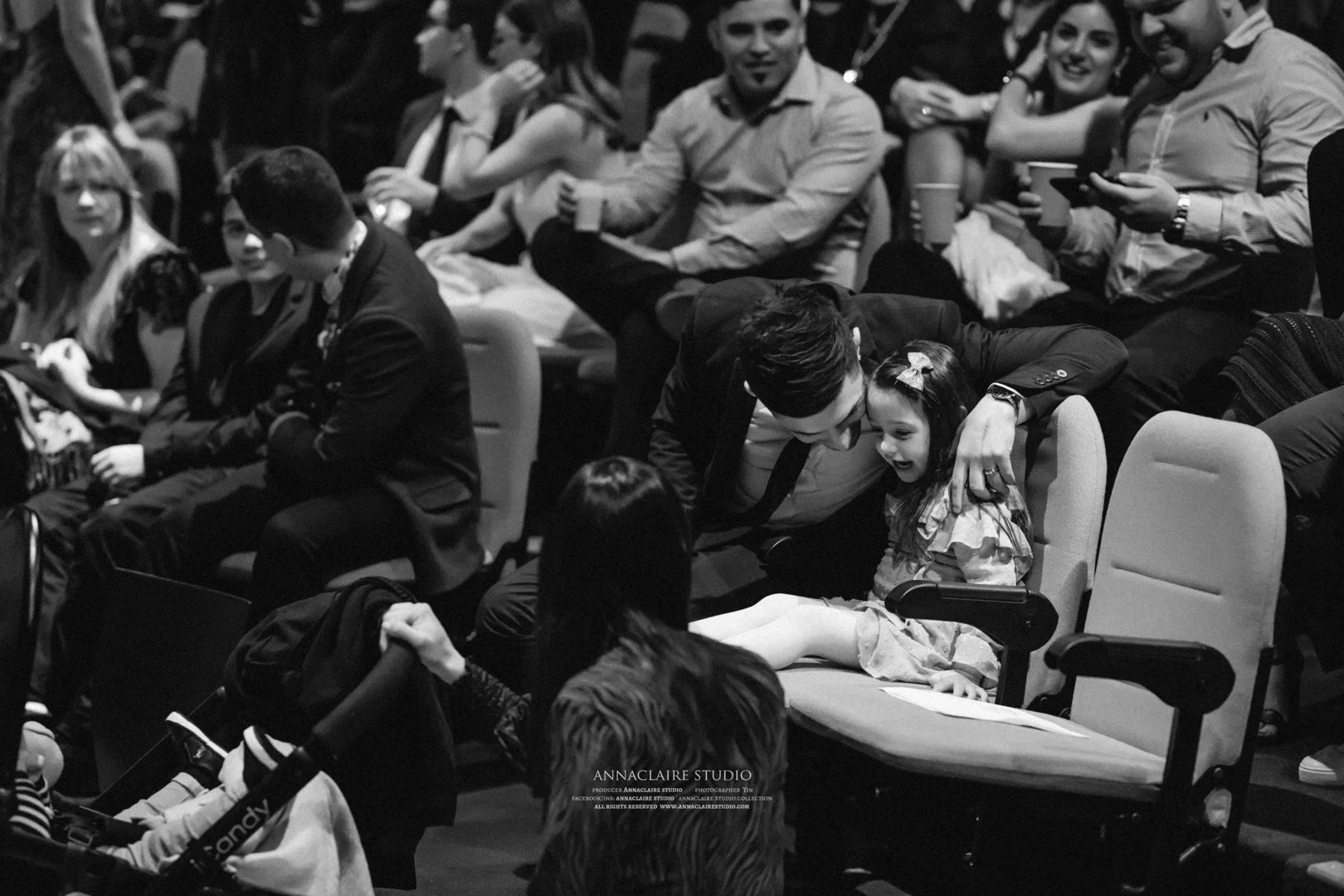A man sitting in a wheelchair interacts with a young girl sitting on a seat, surrounded by people in an auditorium or theater setting.