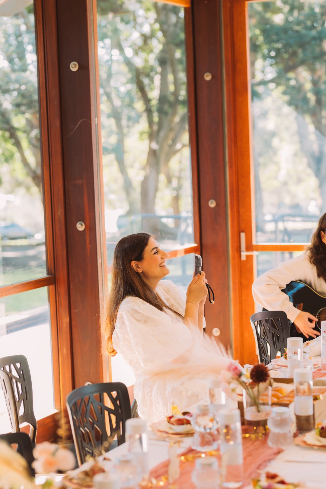 A woman in a white dress sitting at a decorated table, smiling and taking a photo with her phone during a daytime gathering at a venue with large windows and trees outside.