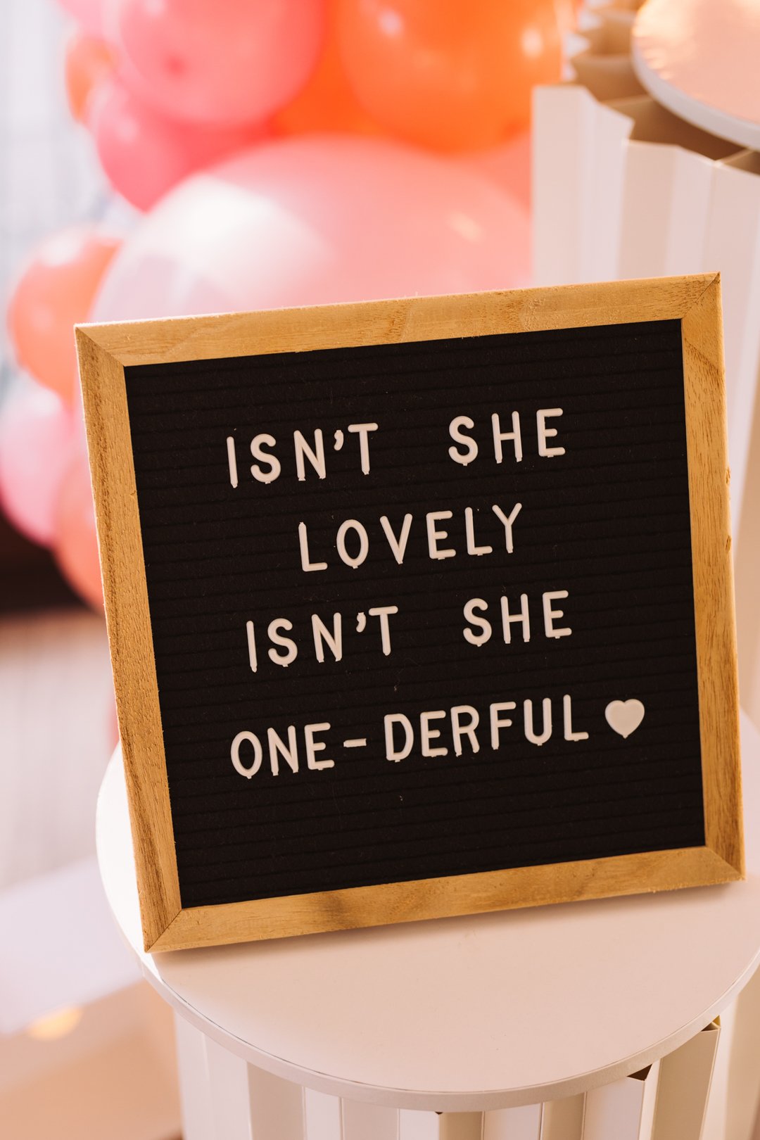 A black letter board with a wooden frame displaying the message: "Isn't she lovely isn't she one-derful" with a small heart symbol, placed on a round white table. In the background, pink and peach balloons are visible.