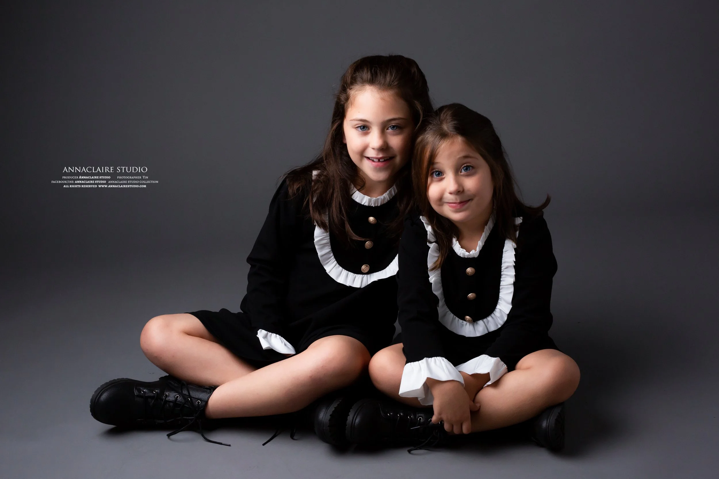 Two young girls sitting cross-legged on grey floor, wearing black dresses with white ruffled collars and buttons, smiling at camera against a dark grey background.