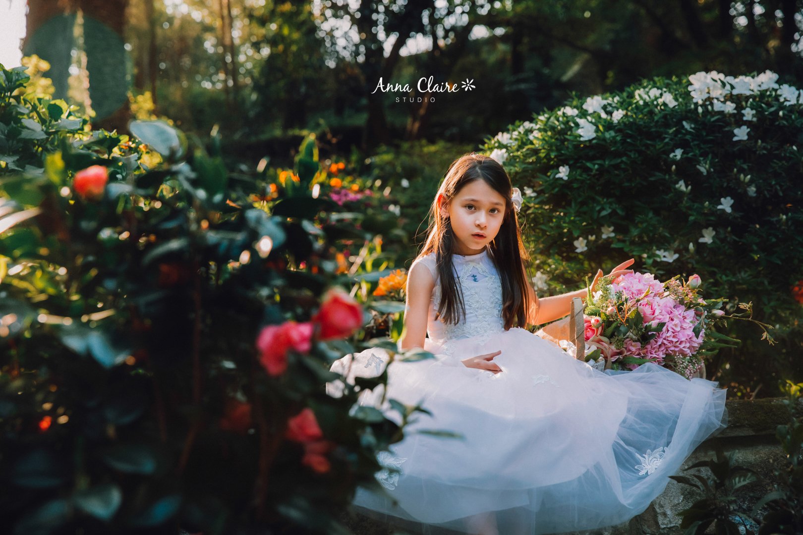 Young girl in a white dress sitting outdoors among green bushes and colorful flowers, holding a bouquet of pink flowers.