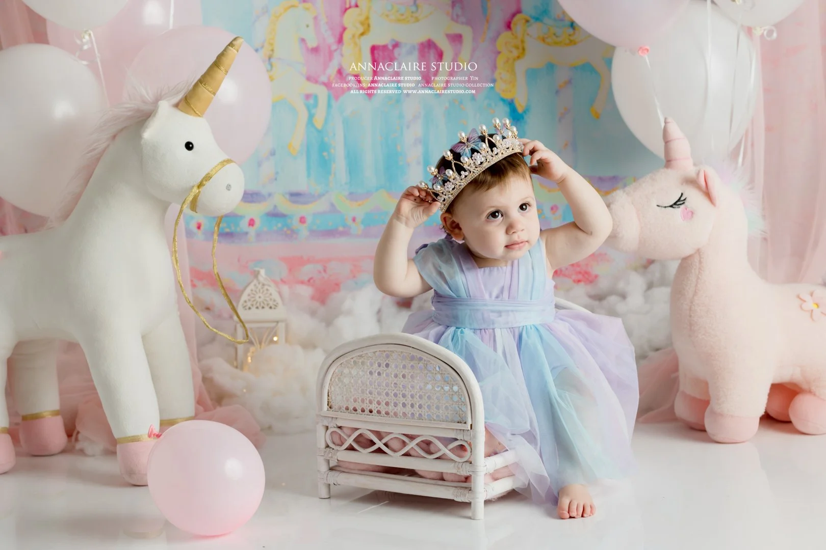 A  one year old baby girl wearing a pastel-colored dress and a princess crown, sitting next to plush unicorns and balloons, with a colorful unicorn-themed backdrop.