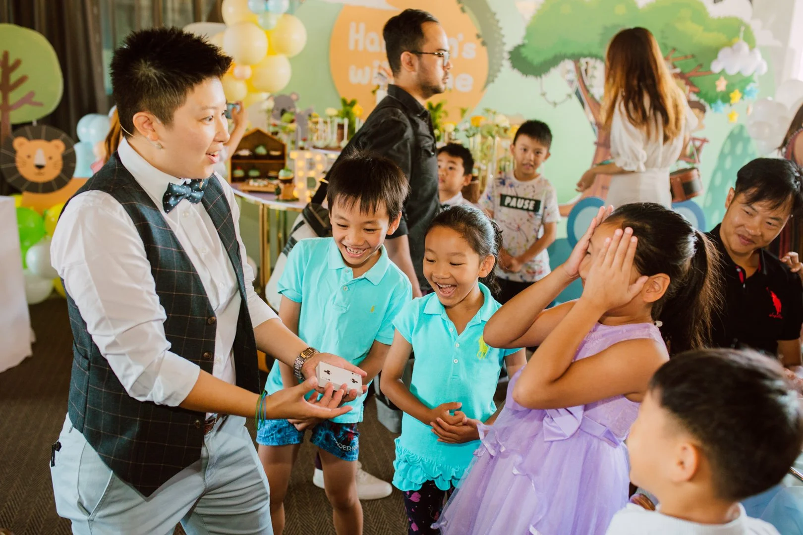 Children and adults at a birthday party, with a boy performing a magic trick for a girl who is surprised, in a decorated room with balloons and colorful backdrop.