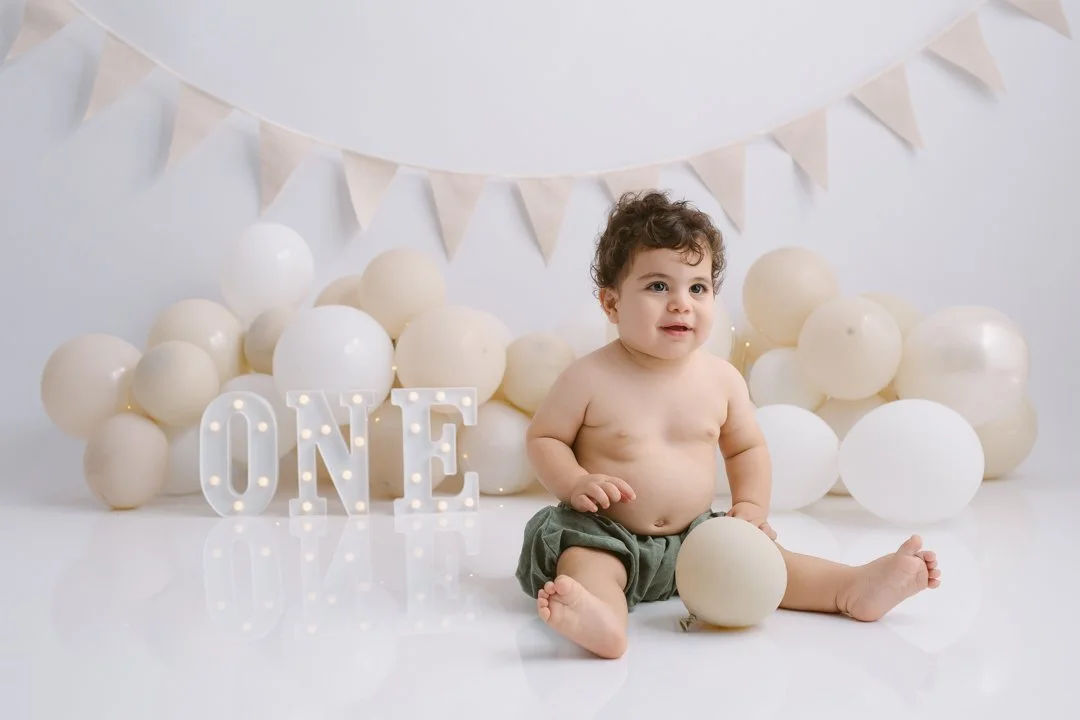 Baby sitting on the floor surrounded by white balloons, with a lit-up letter sign 'ONE' behind him, beige bunting hanging above.