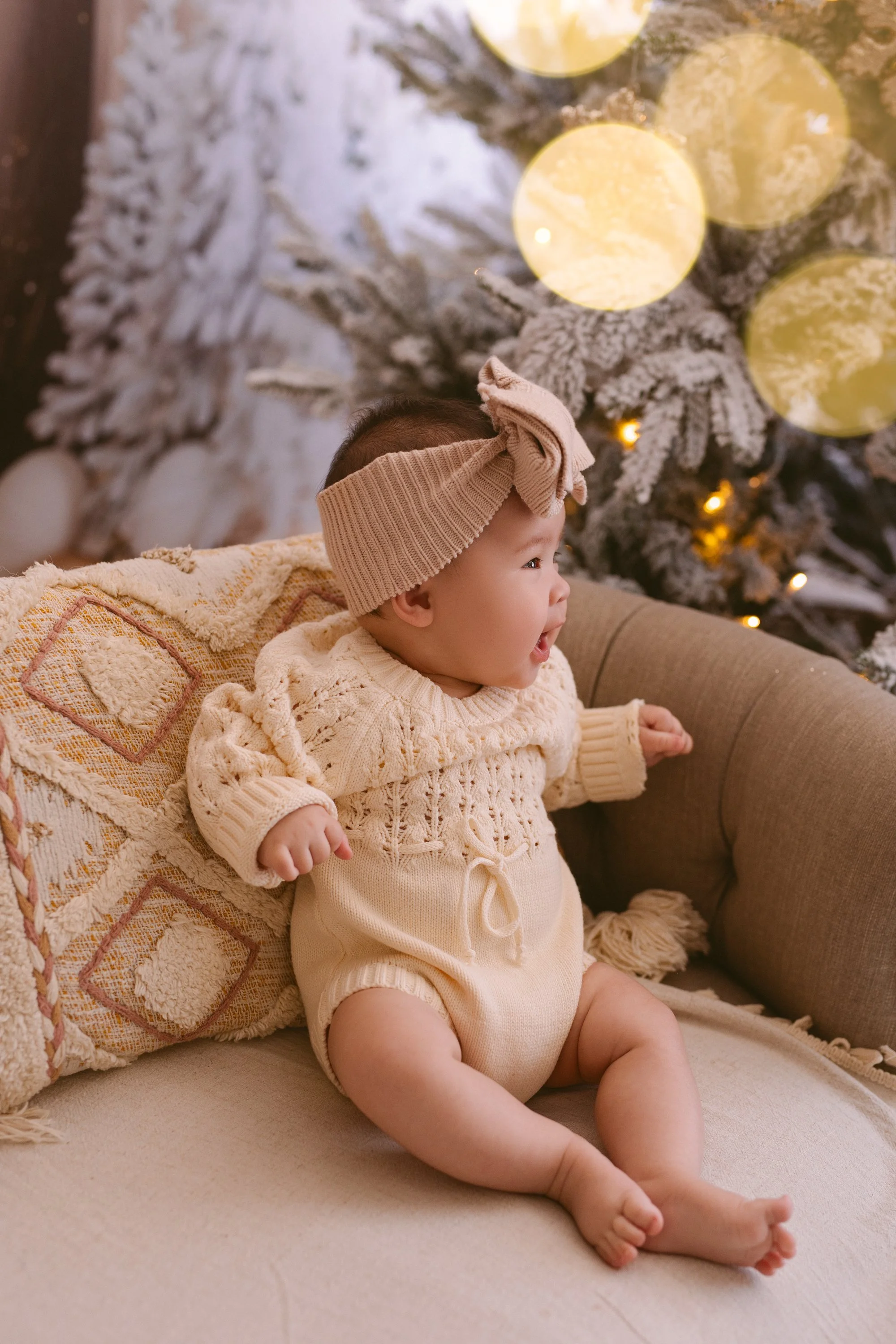 A happy baby sitting on a beige couch with a decorative pillow, wearing a knit sweater and a beige headband with a bow, in front of a snow-covered Christmas tree with glowing lights.