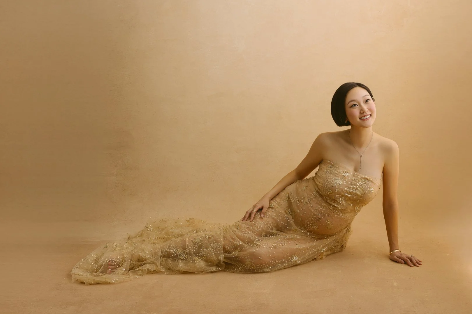 A woman in a gold, sequined dress lying on the floor against a gold background, smiling and looking to her left.