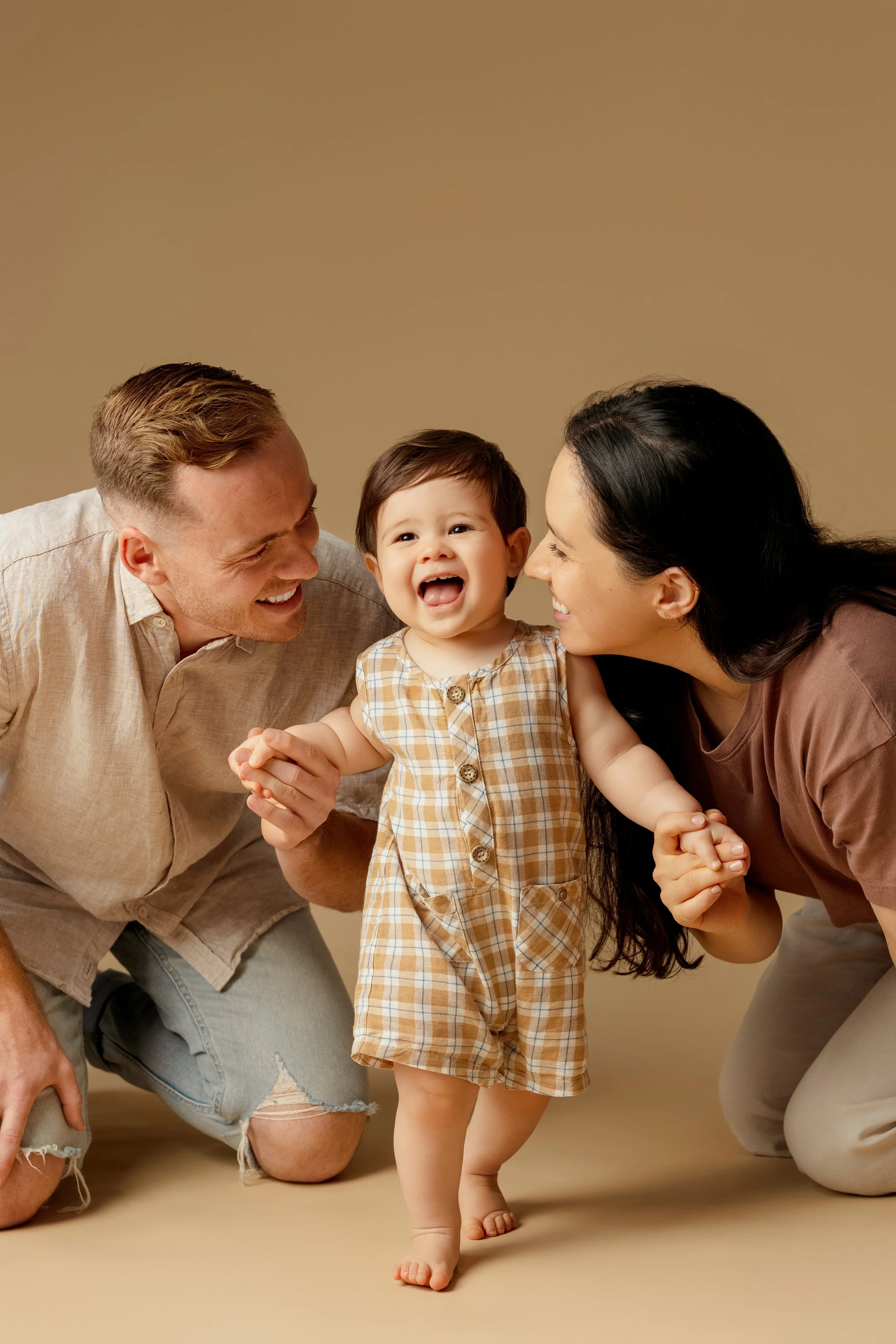 A happy toddler walking while holding hands with a man and woman, likely parents, on a beige background.