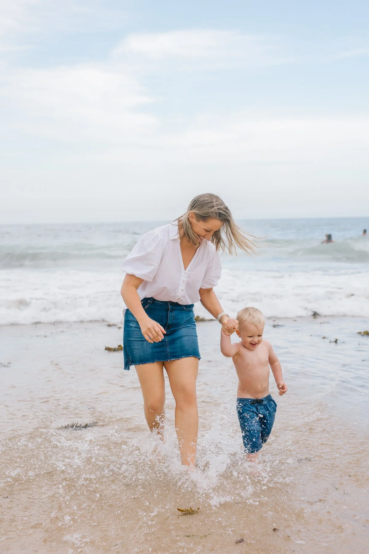 A woman and a young boy are walking hand in hand at the beach, splashing in shallow water with waves in the background.