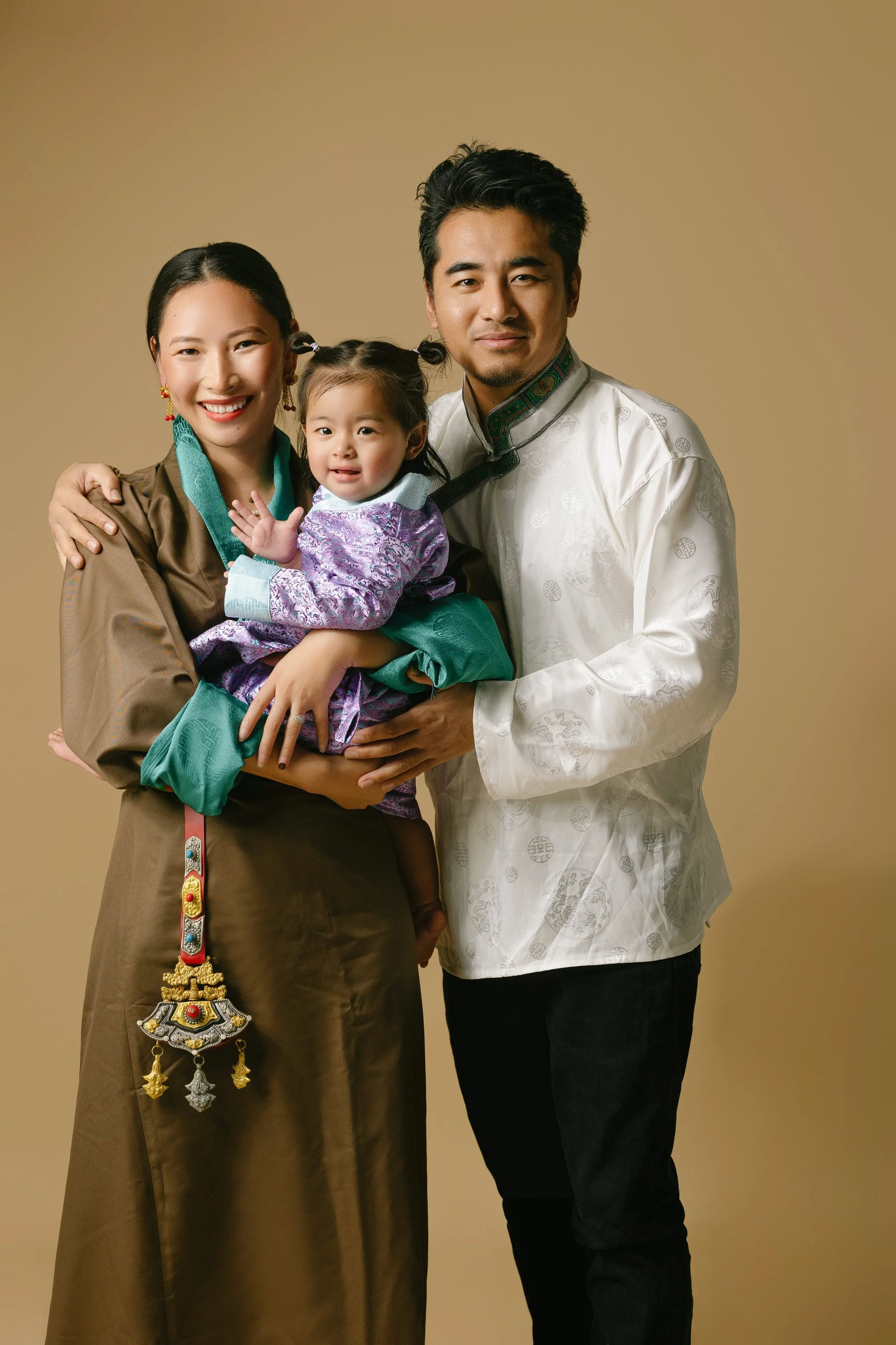 A family of three dressed in traditional Asian clothing, standing together against a plain beige background. The woman is holding a young girl, and a man is standing beside them.