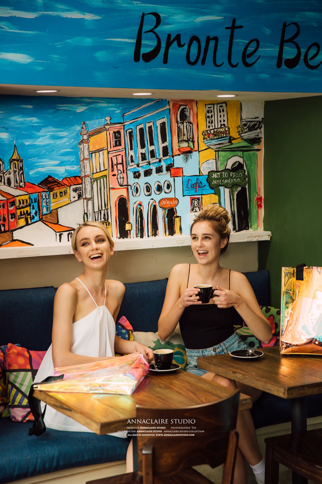 Two young women sitting at a table in a colorful cafe, smiling and holding cups of coffee, with a vibrant mural of a cityscape in the background.