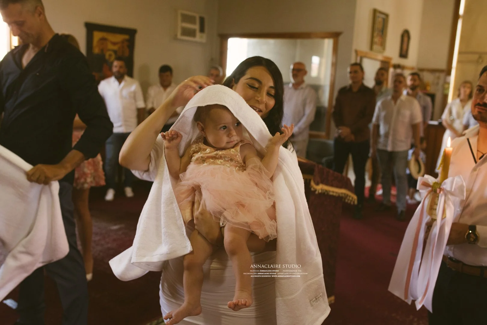 A woman holding a young girl dressed in a peach-colored dress with a towel over her head, in a room with several people watching, likely at a celebration or religious ceremony.