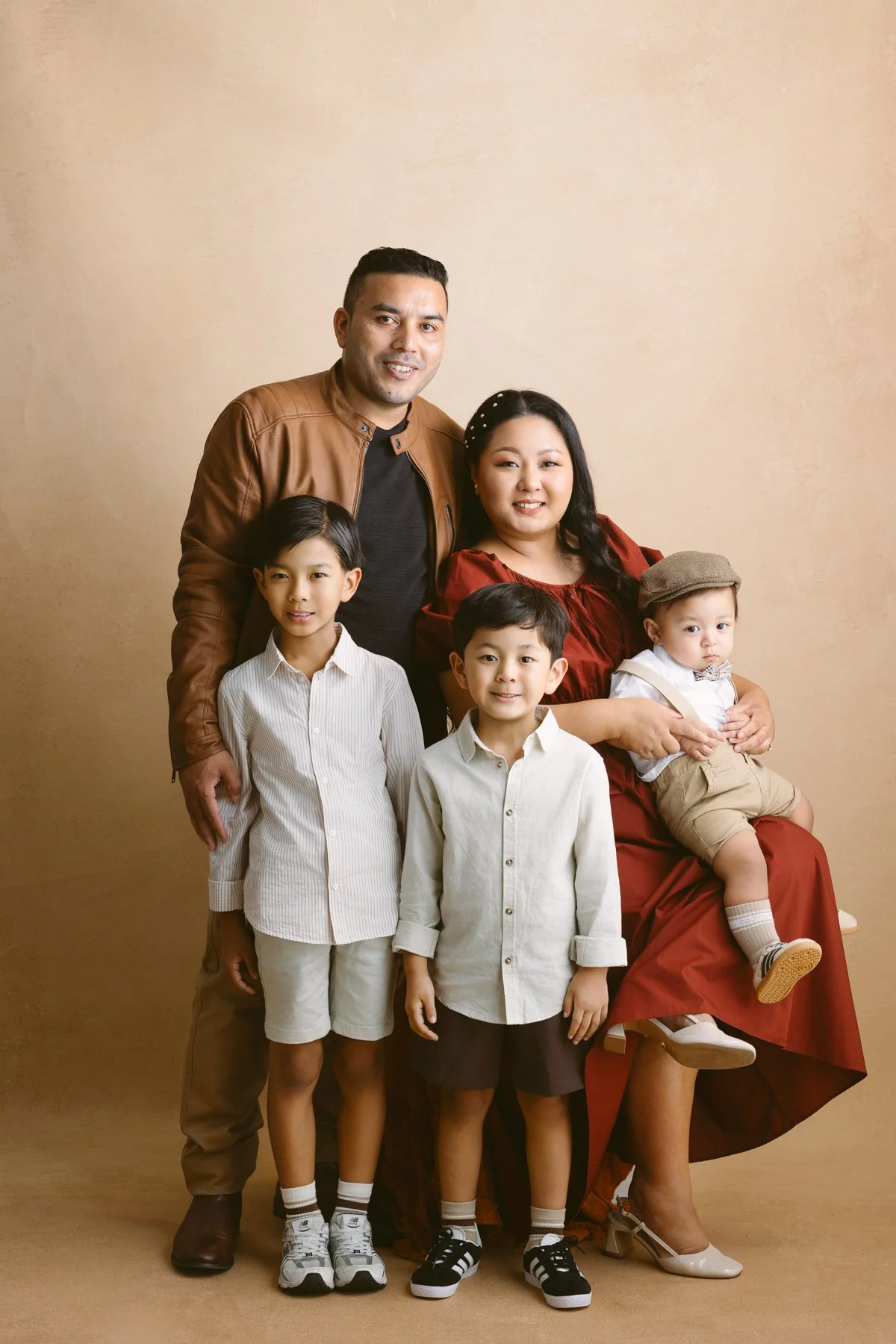 A family of six posing for a photo against a beige background. The father, in a brown jacket, and the mother, in a red dress, are standing behind four children. The children, two boys in white shirts, one boy in brown shorts, and a toddler girl in be