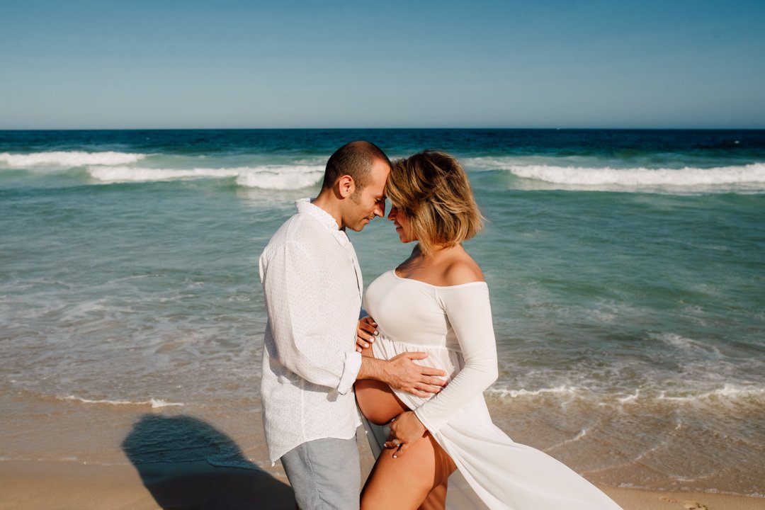 mum to be and husband standing close on the beach, touching foreheads with shadows cast on the sand, ocean waves in the background.