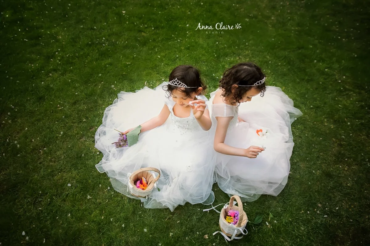 Two young girls in white dresses sitting on grass, holding flowers and surrounded by small baskets of flowers, wearing tiaras, with a lush green lawn background.