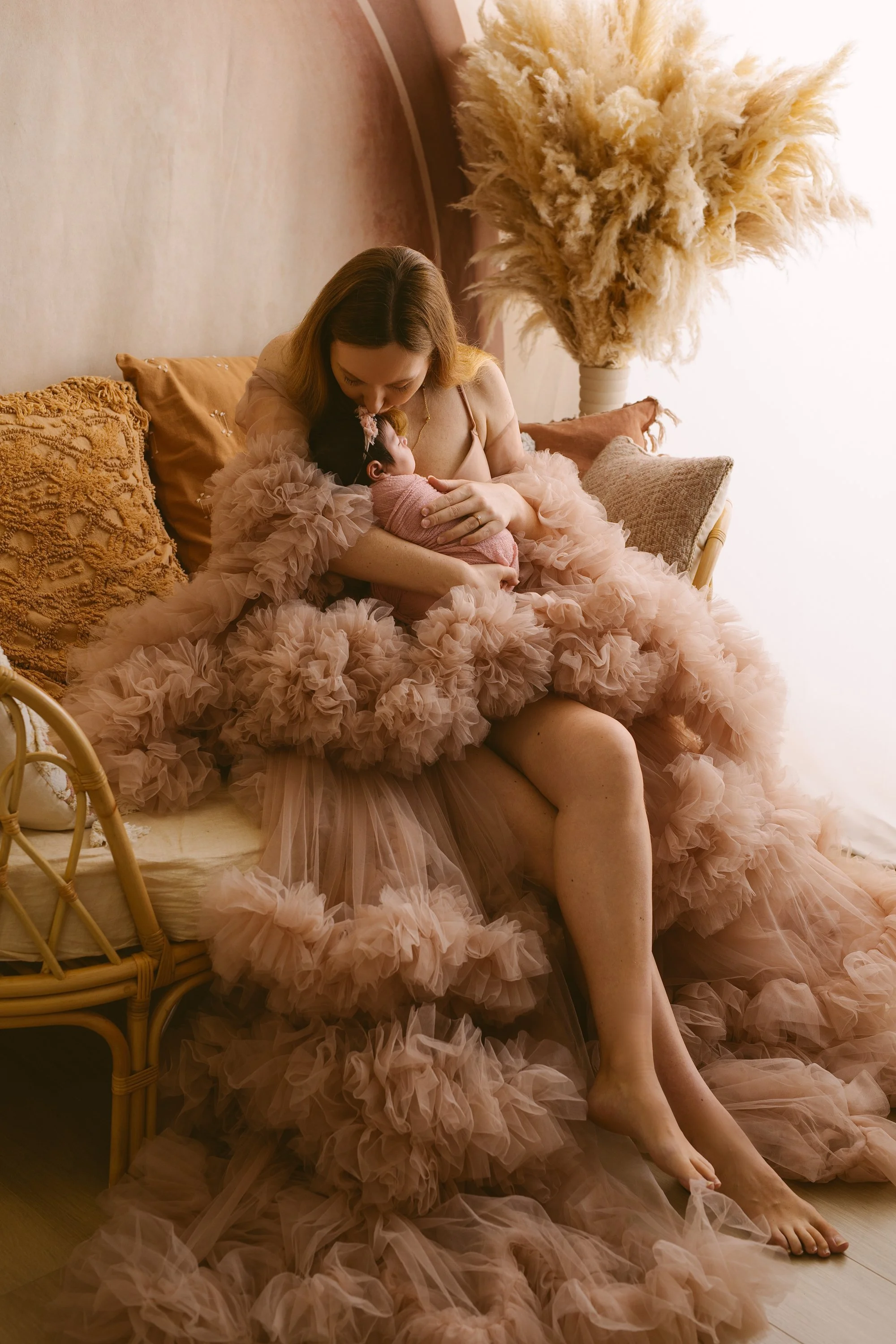 A mum wearing a peach-colored ruffled gown sitting on a sofa and breastfeeding a newborn baby in a pink outfit, in a warmly lit room with a large feathered plant in the background.