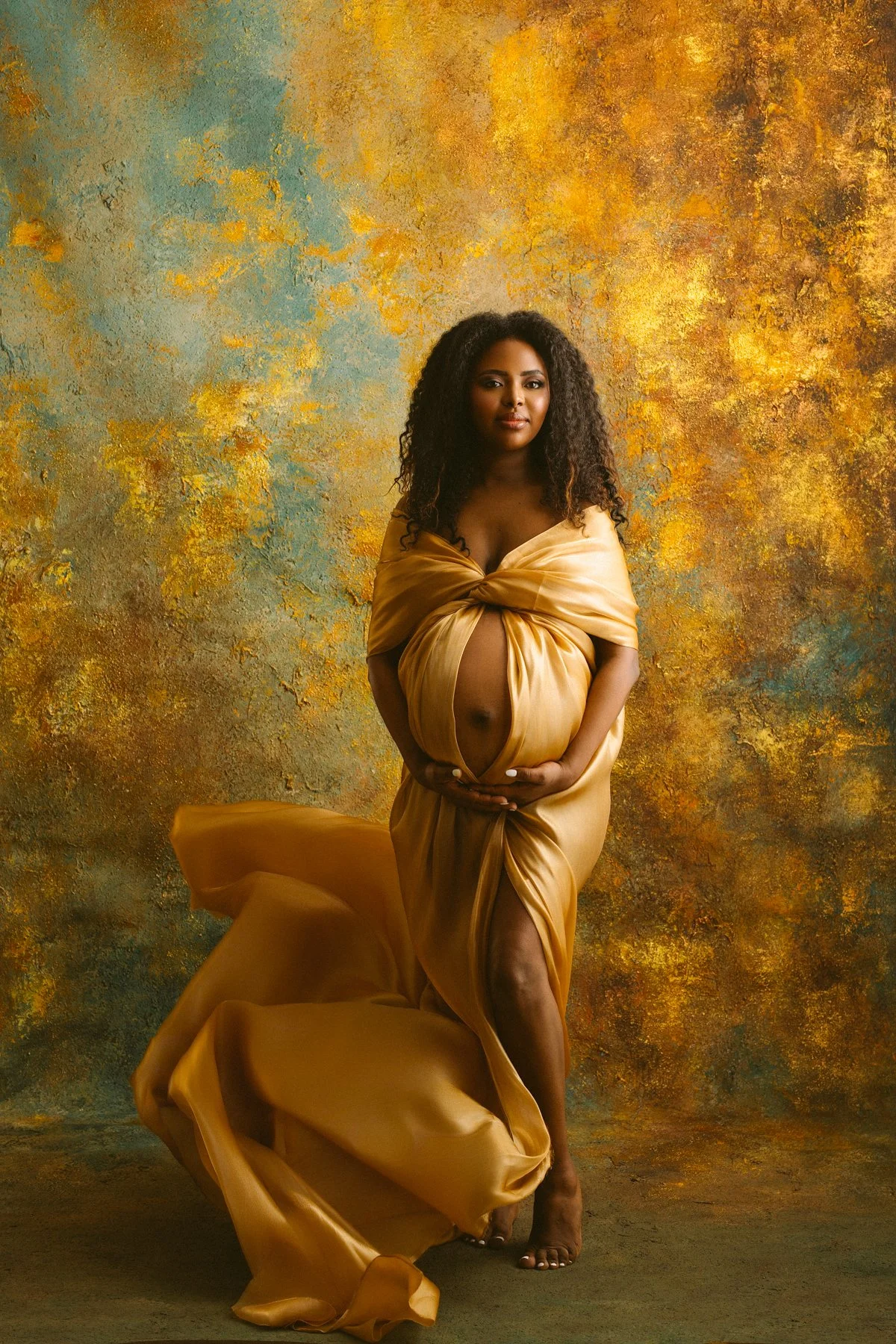 A pregnant woman in a flowing gold dress stands against a textured yellow and green background, holding her belly with both hands.