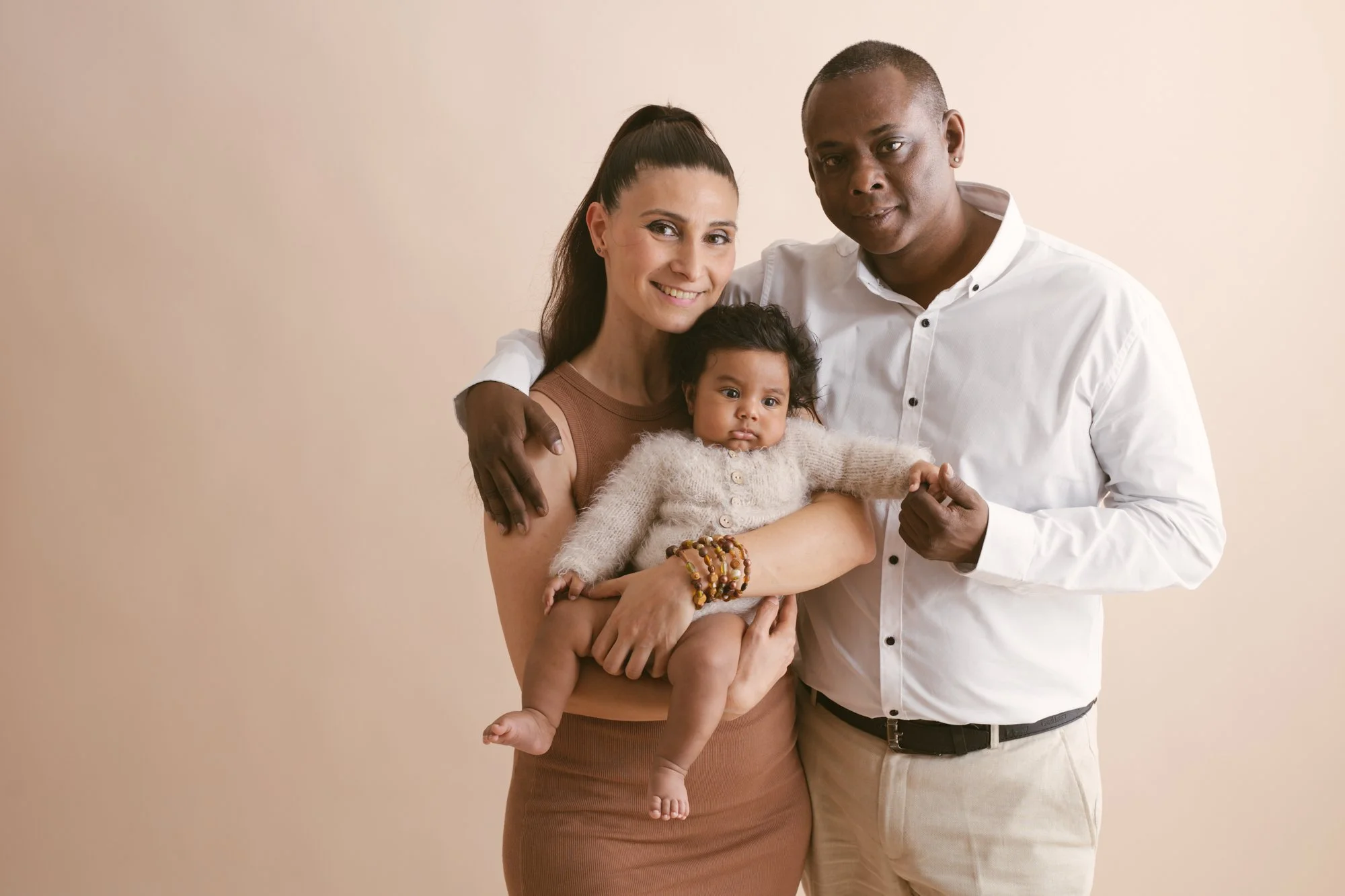 A happy family of three standing together against a neutral background, with a woman holding a young child and man standing beside them, all looking at the camera.