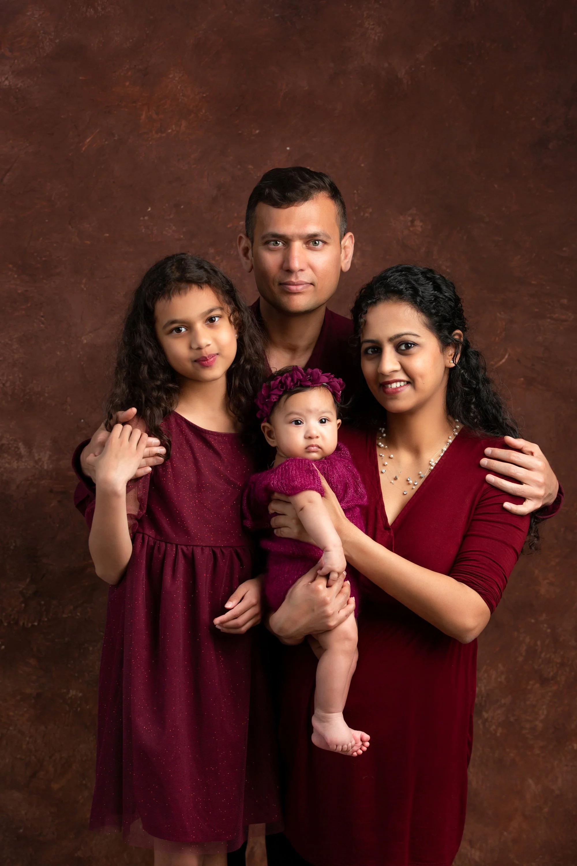 A family portrait featuring a man, woman, and two young girls, all dressed in matching burgundy outfits, standing against a brown textured background.