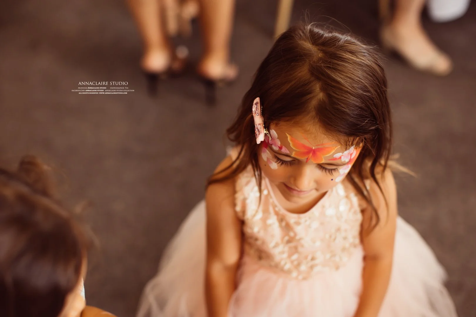 A young girl with long brown hair and face paint of a butterfly and flowers, wearing a light-colored dress with lace details, is looking downward with her eyes closed, surrounded by other children at what appears to be a social gathering or party.