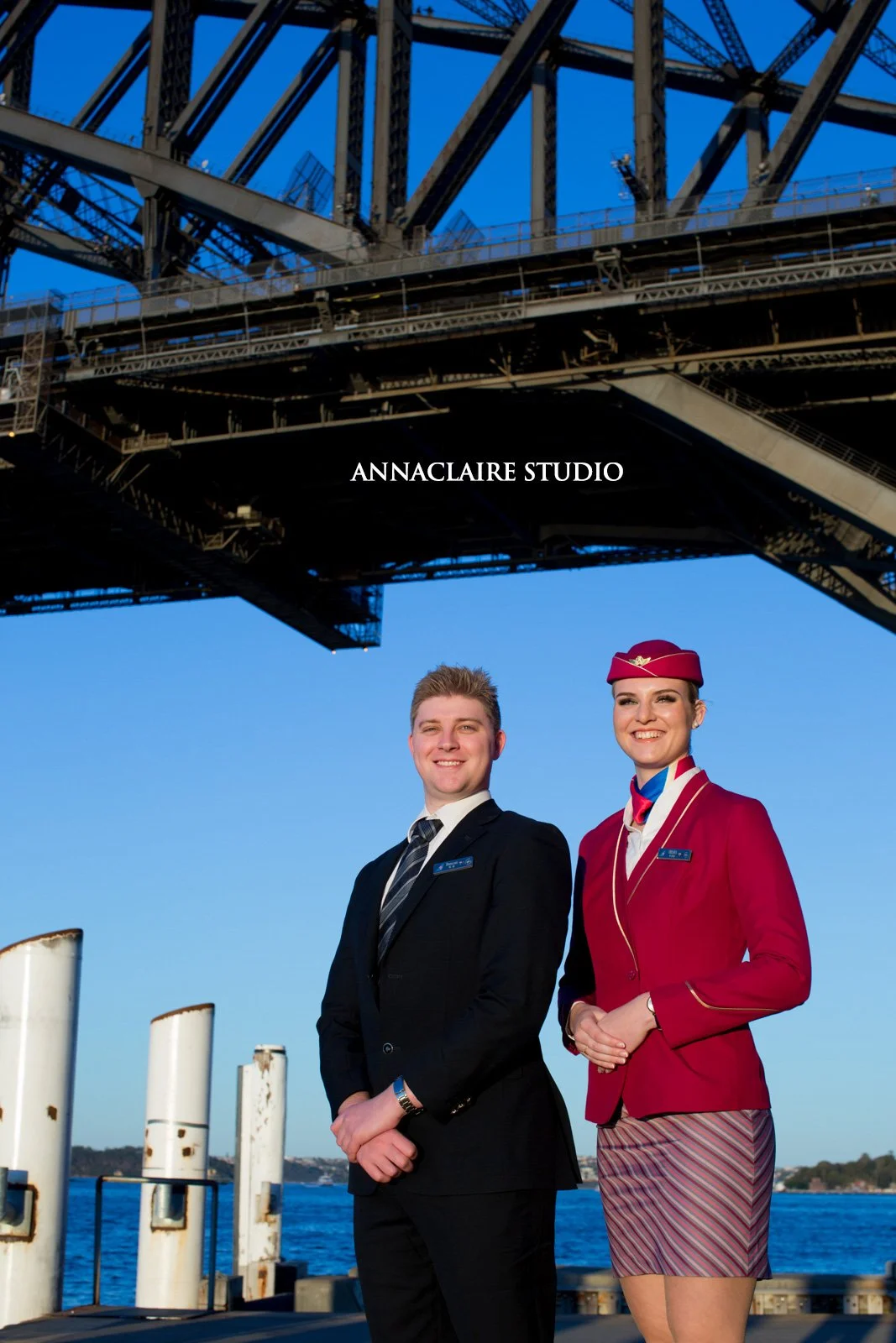 A man and a woman dressed as airline crew standing under a bridge over water, smiling at the camera.