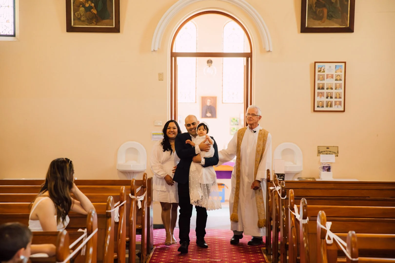 A family standing inside a church during a religious ceremony. A man in a suit is holding a young girl in a white dress, while a woman and an elderly man stand beside them. The church has wooden pews, religious artwork on the walls, and stained glass