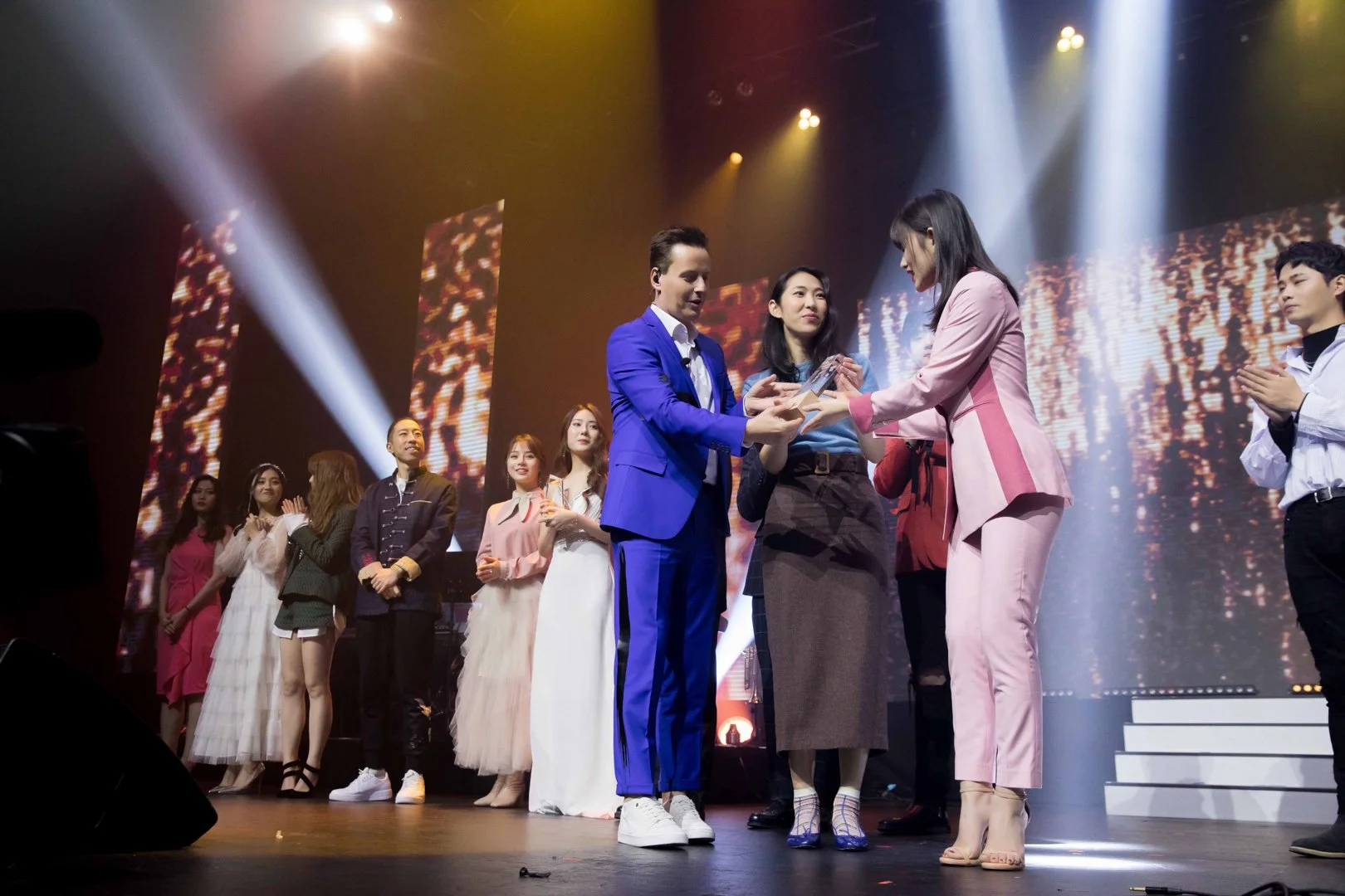 Group of people on stage during an award or recognition ceremony, with two women in pink suits handing an award to a man in a blue suit, while others stand in the background watching.