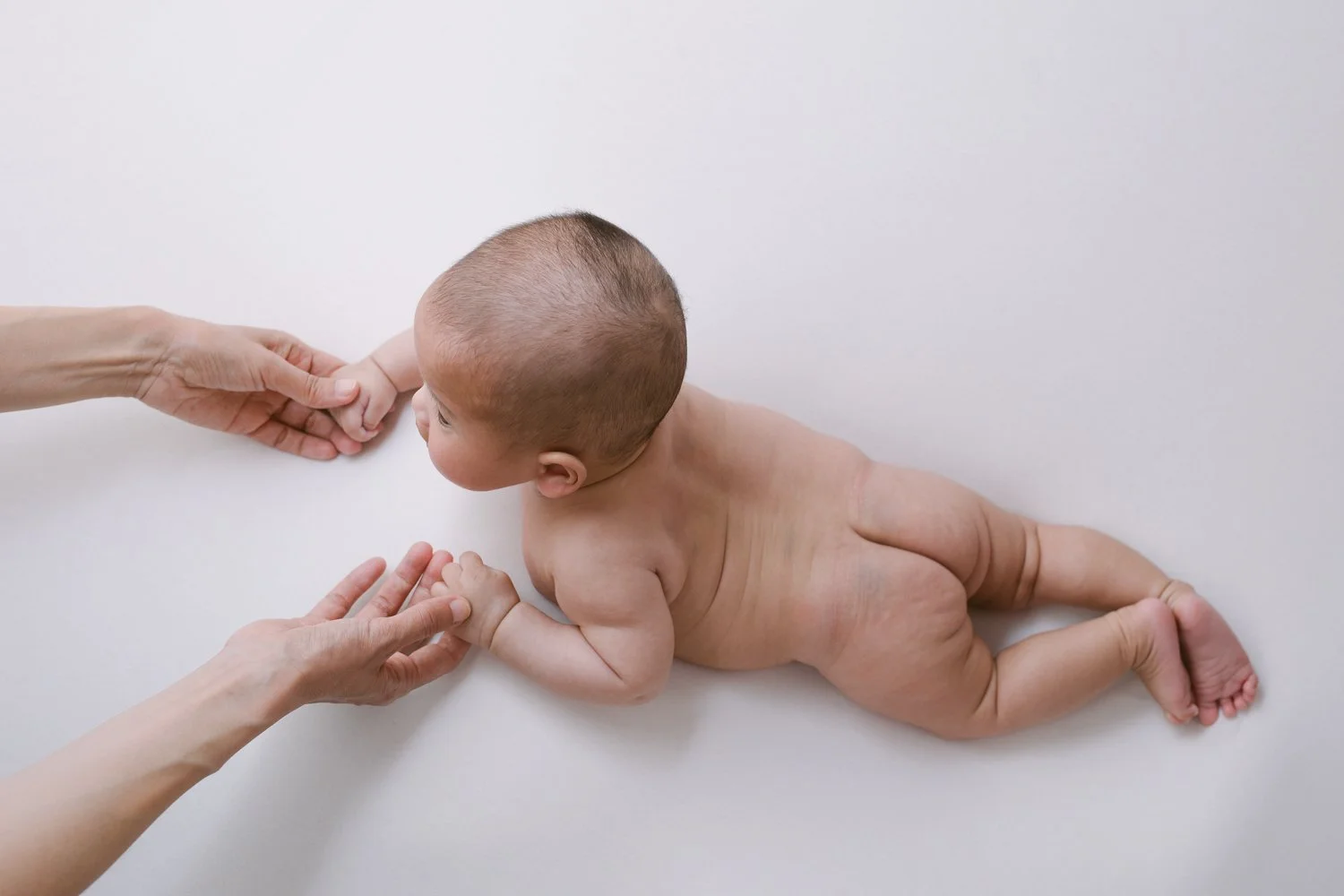 A naked baby laying on a white surface, holding hands with two adults, one on each side, in a tender moment.