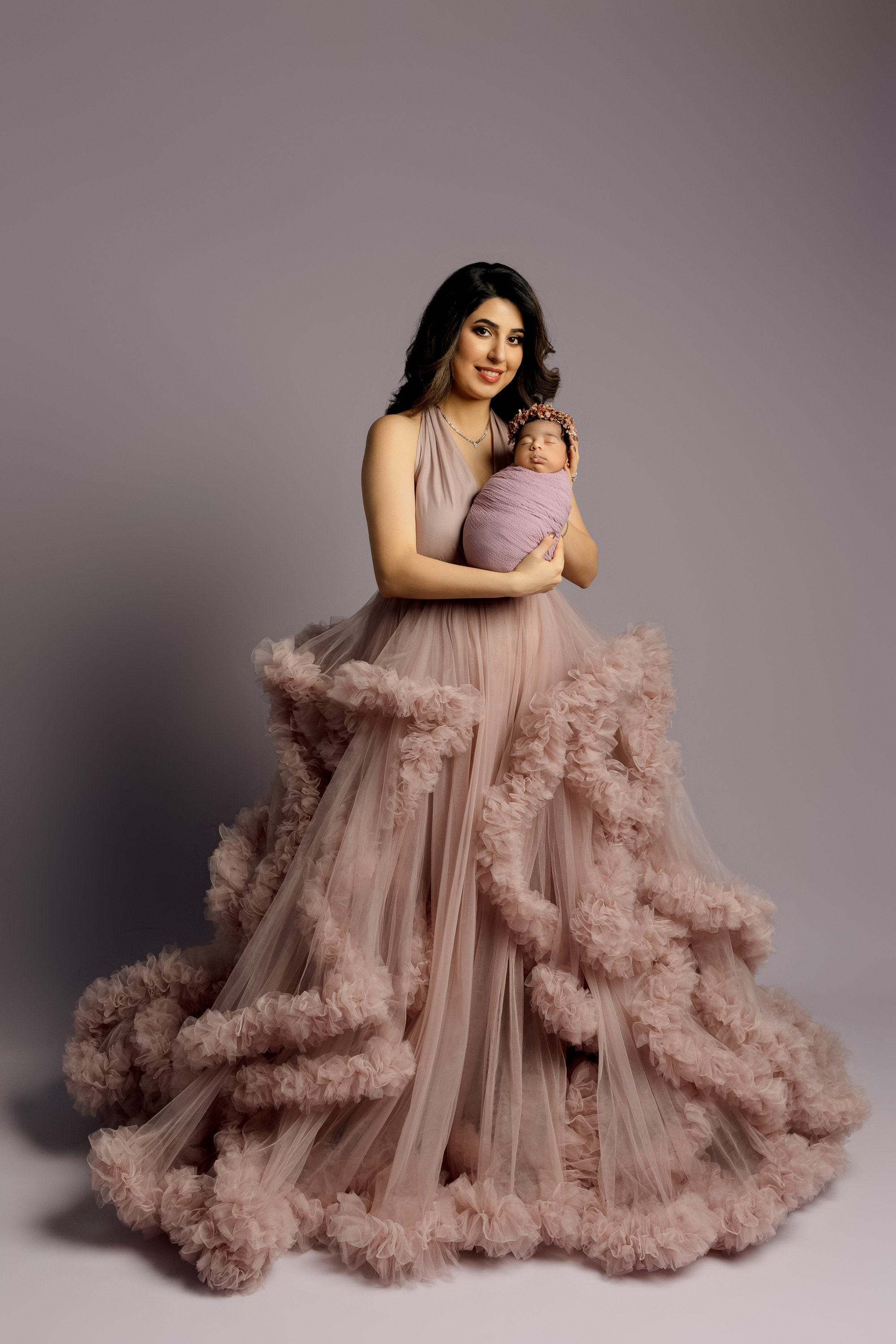 A woman in a pink, ruffled, tulle gown holding a newborn wrapped in a lavender blanket with a matching headband, standing against a plain light gray background.