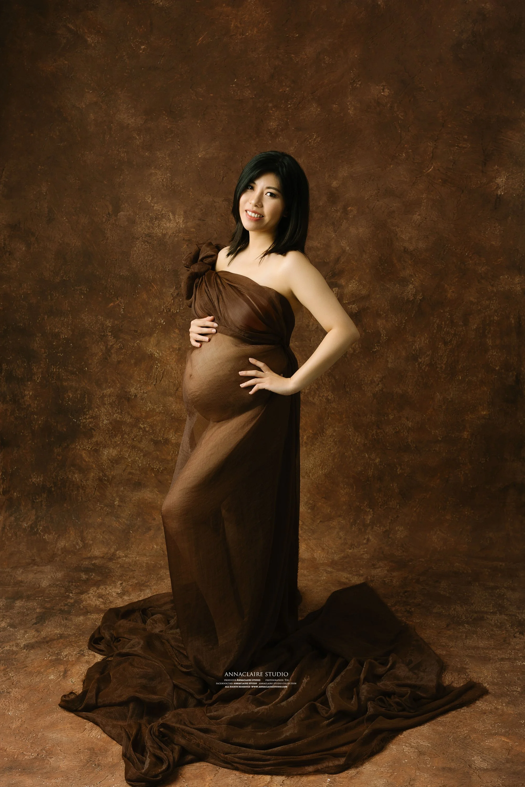 Pregnant woman in a brown dress standing against a brown textured background, smiling with one hand on her belly and one on her hip.
