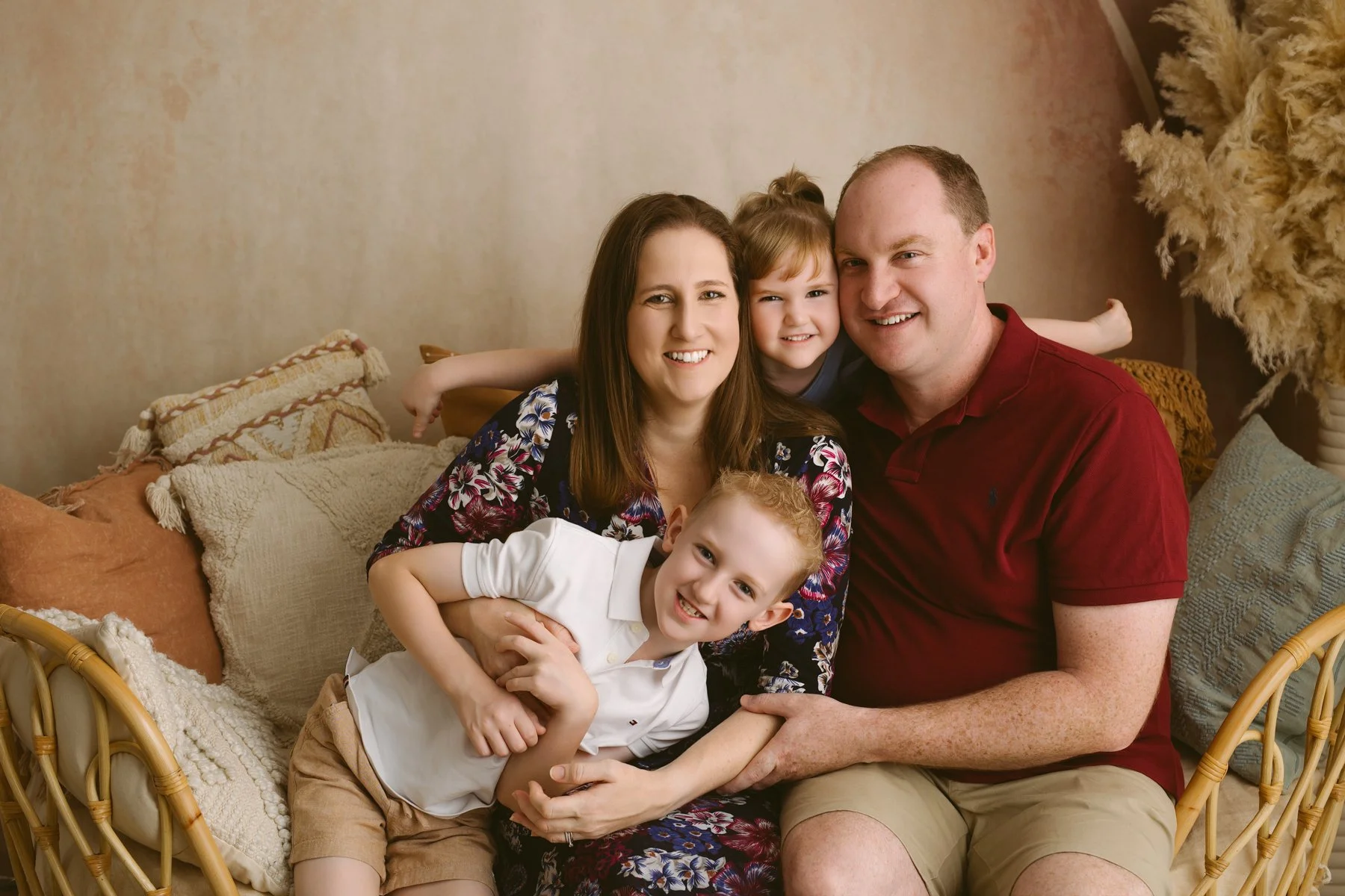 A smiling family of five sitting on a beige couch, with mum ,dad , and 2 children, all close together in a cozy living room.