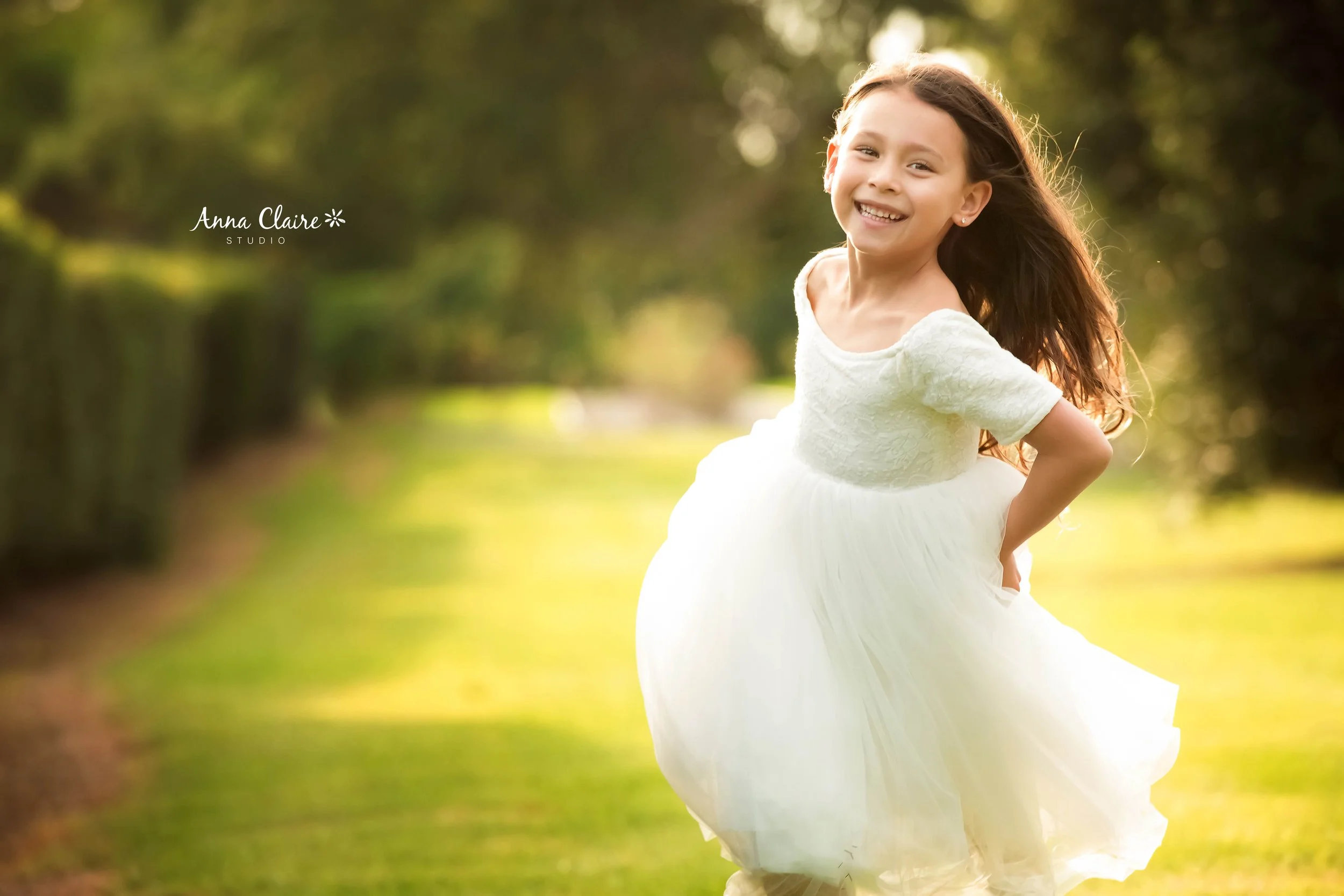 A young girl in a white dress laughing outdoors in a sunny park with green trees and a grassy pathway.
