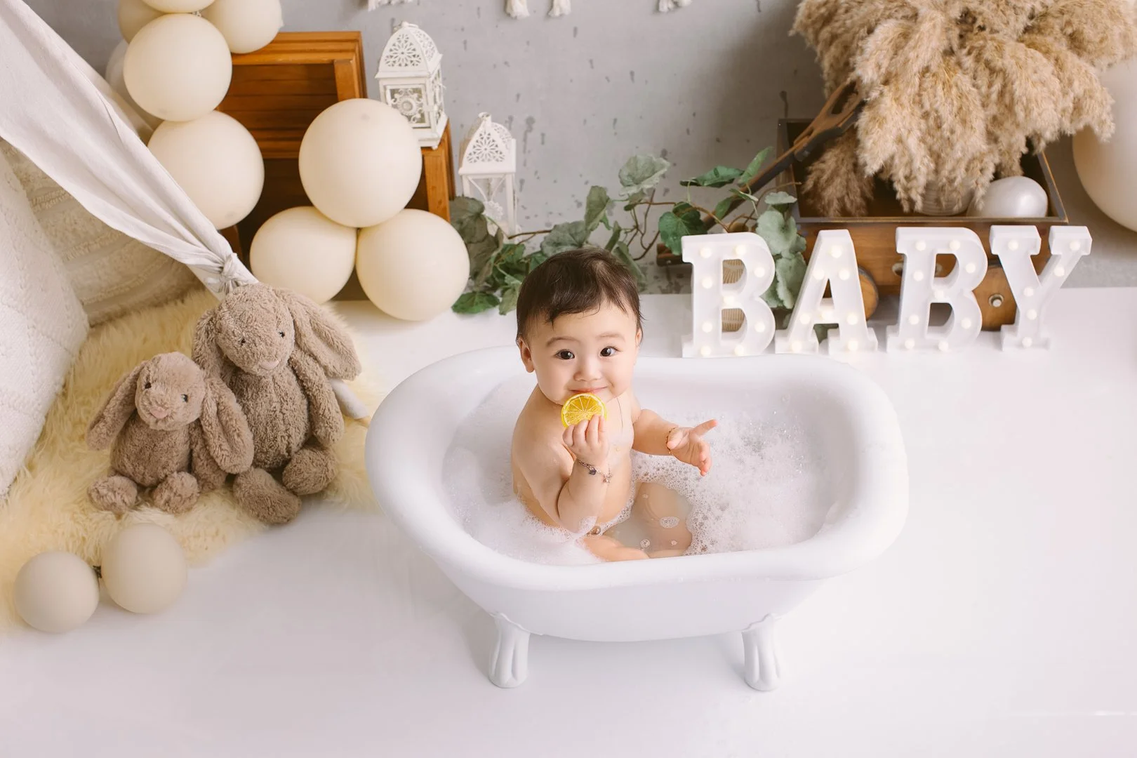 A  one year old girl sitting in a white bathtub filled with bubbles, holding a slice of lemon and smiling.