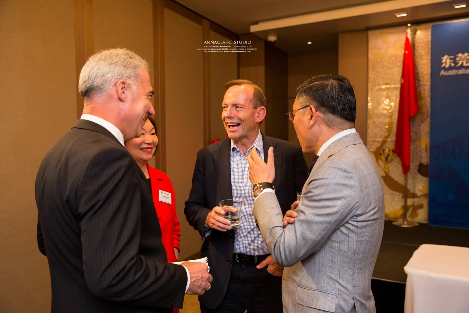 Four people, three men and one woman, are engaged in conversation and smiling at a professional event. The woman is wearing a red blazer, and the men are dressed in suits. One of the men is holding a glass of water.