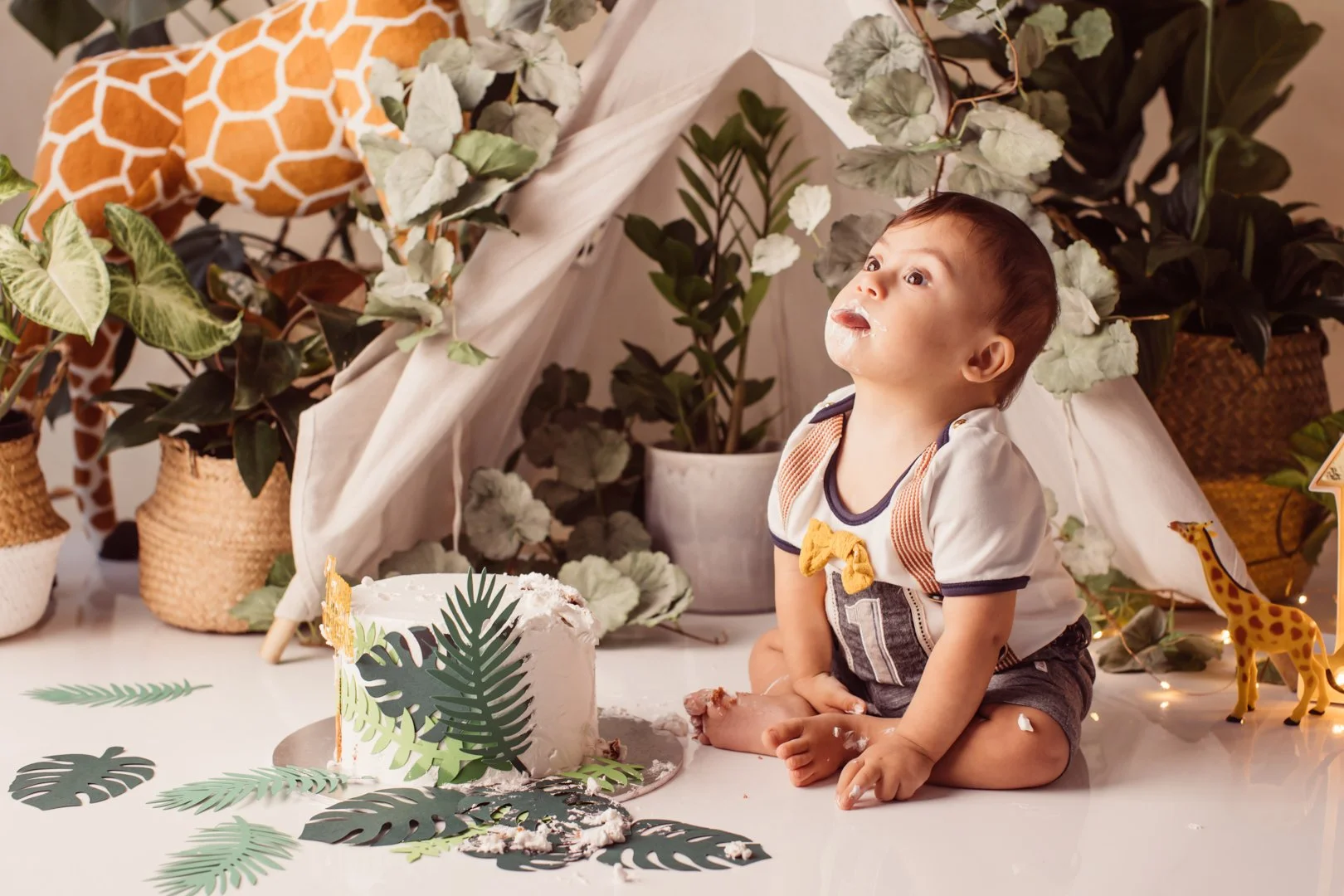 1 yr old boy  with a messy face sitting on a floor next to a small cake decorated with tropical leaves, surrounded by greenery and jungle-themed cake smash photo decorations.