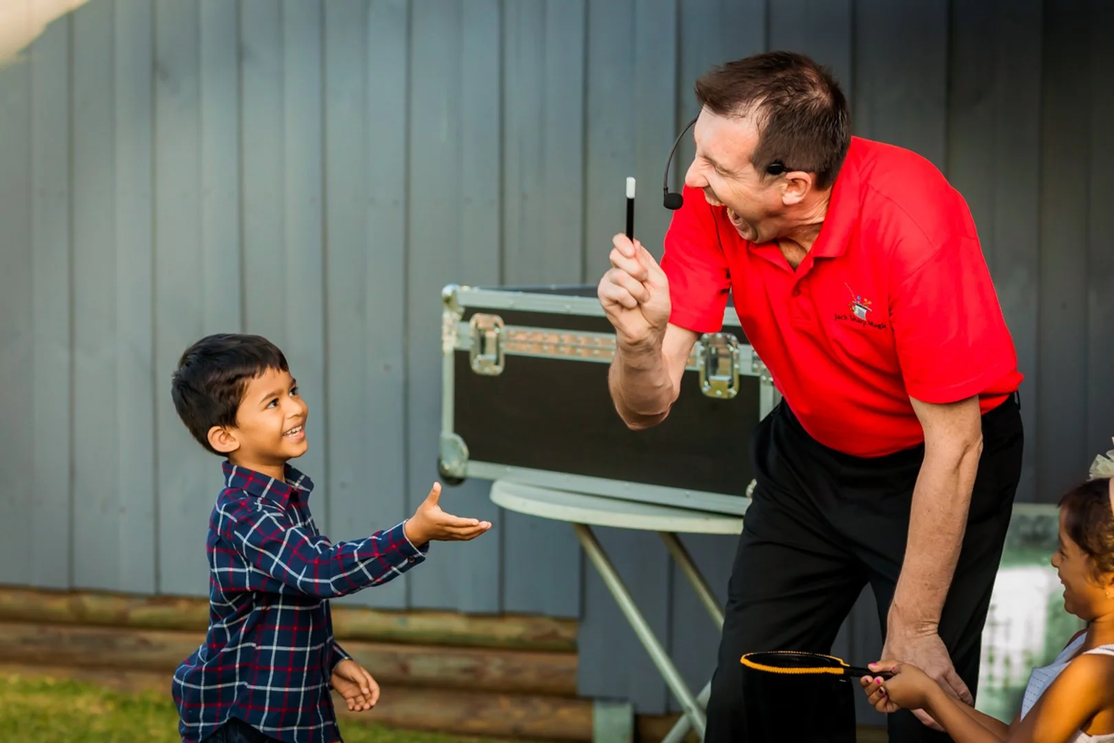 An adult man in a red polo shirt holding a microphone, laughing and pointing. A young boy in a plaid shirt smiling and reaching out. A young girl holding a tennis racket, smiling. They are outside near a blue wall and a table with a box.