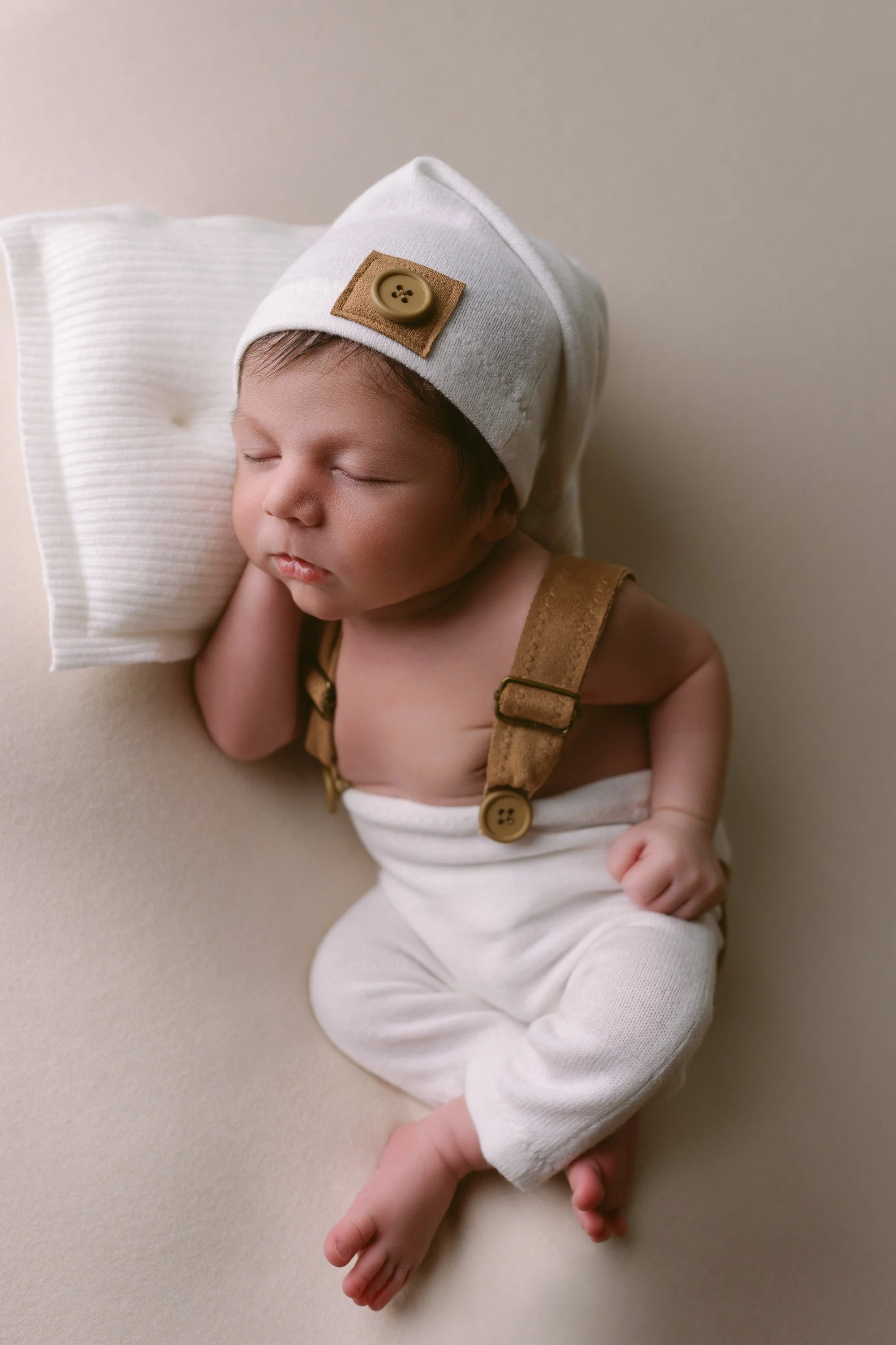 A sleeping newborn baby with a white hat decorated with a button, lying on a cream-colored surface, wearing white pants and suspenders.
