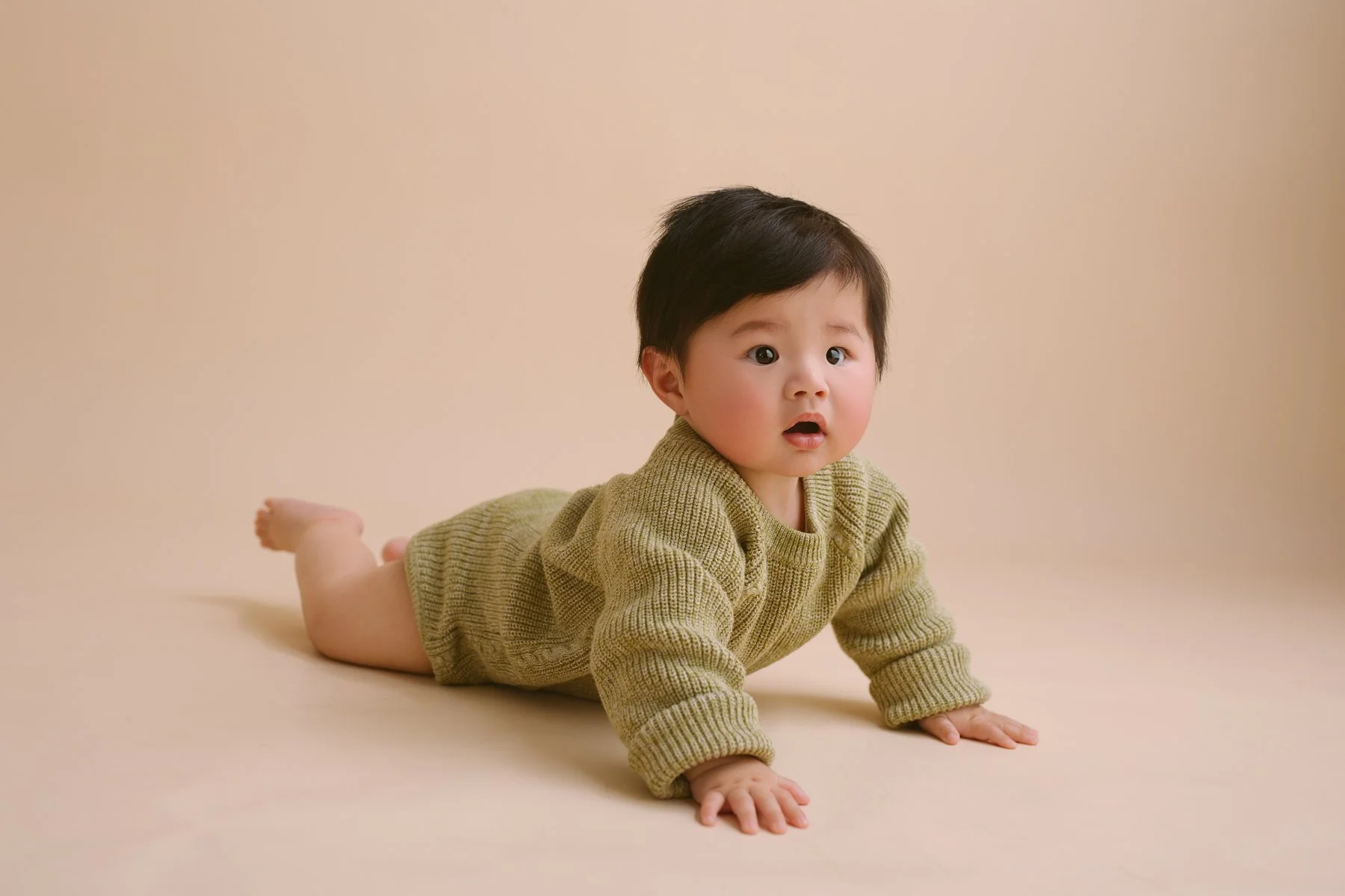 Young child crawling on a beige surface, wearing a green knitted sweater.