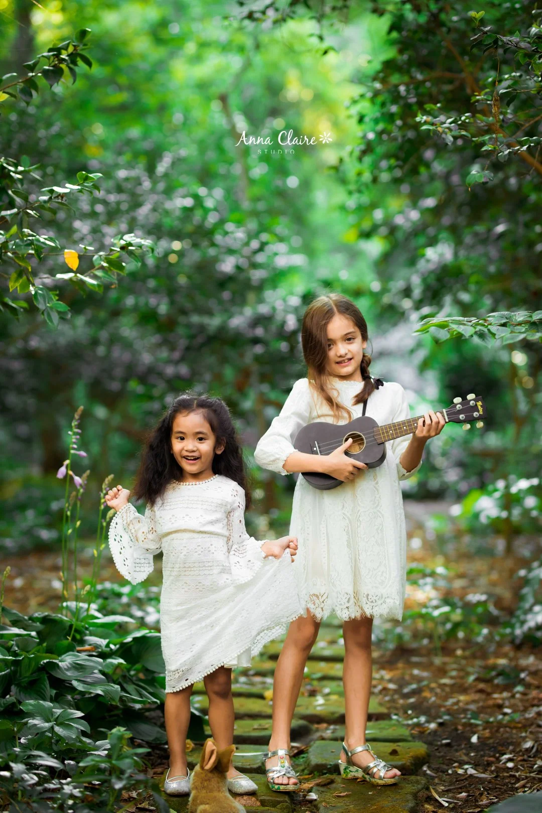 Two young girls in white dresses standing on a wooded trail. The older girl is playing a small black guitar, while the younger girl is holding her dress and smiling. Lush green foliage surrounds them in a natural outdoor setting.