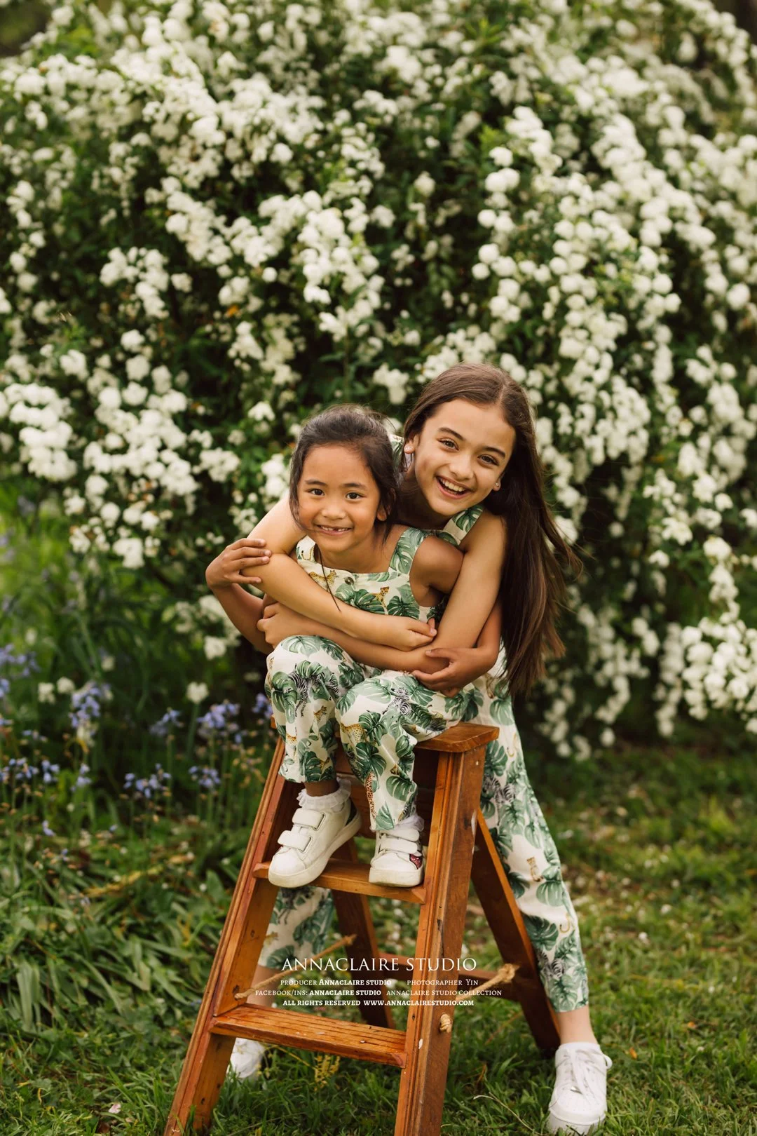 Two girls, one older and one younger, sitting on a wooden step ladder outdoors, smiling and hugging each other, with a background of white flowering bushes and green grass, wearing matching tropical-themed outfits.