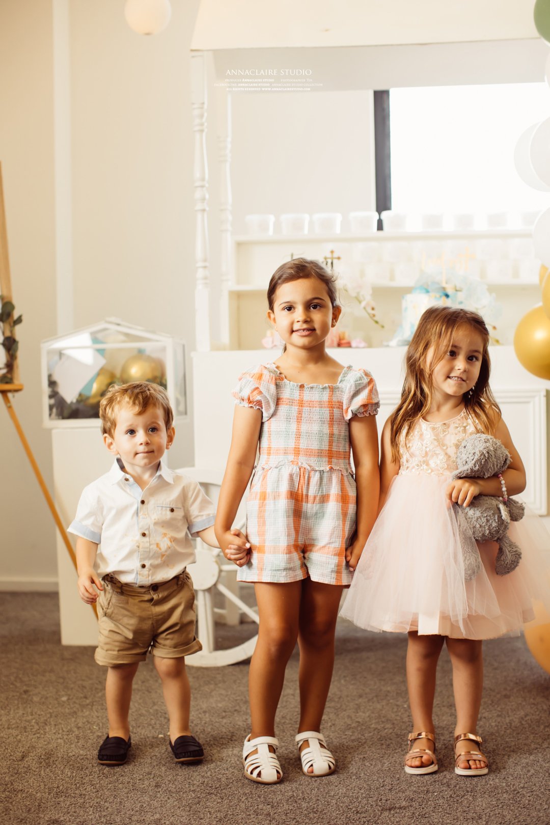 Three children standing indoors at a celebration, holding hands. The boy is on the left wearing a white shirt, khaki shorts, and black shoes. The girl in the middle is wearing a short-sleeved plaid romper and white sandals. The girl on the right is i