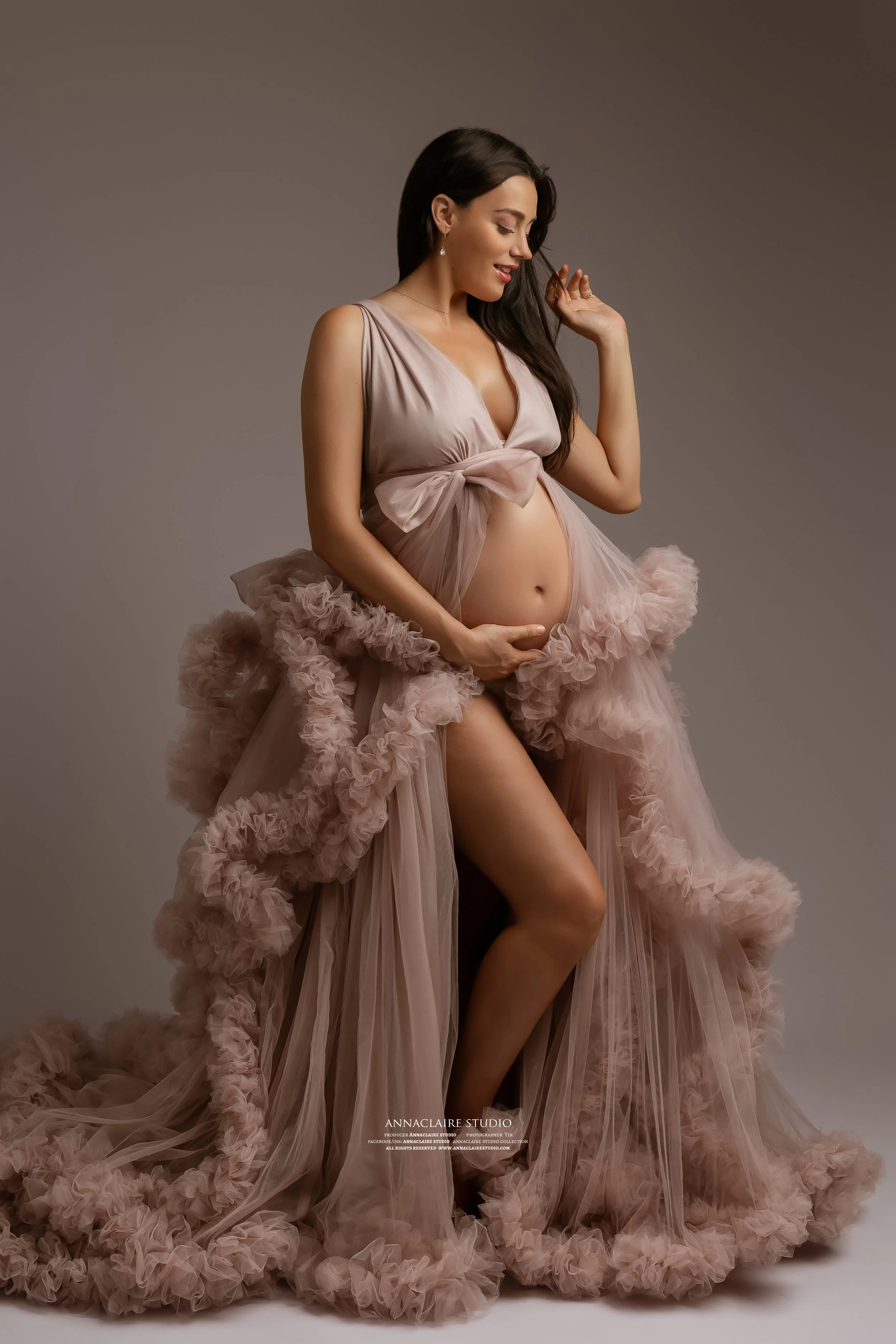 A pregnant woman in a flowy, ruffled pink dress poses for a studio photo, gently holding her belly and smiling.